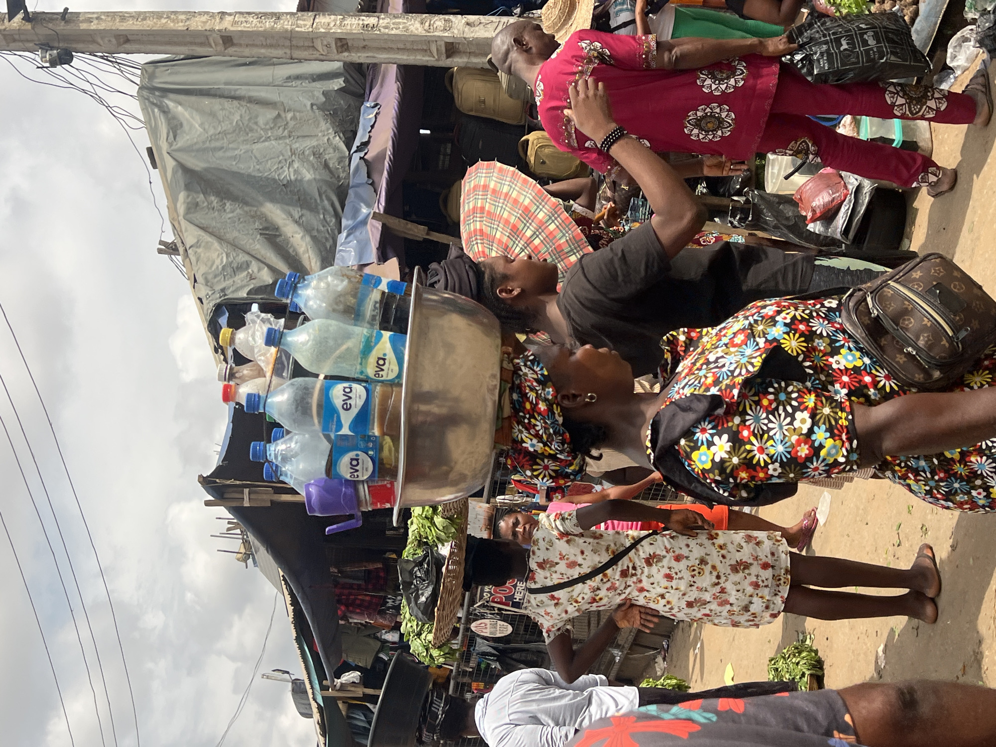 This media file shows an outdoor market scene in Nkpor, Nigeria. The image is oriented vertically, making it appear as if the viewer is looking up.
The primary subjects are several people. In the foreground, a woman wearing a white dress with a floral pattern is visible from the waist down, standing and facing away from the camera. Her legs are bare, and she is wearing sandals. To her left, another person is mostly obscured, but a portion of a person in a white shirt is visible. In the mid-ground, a woman with dark hair, wearing a colorful dress with a floral print, is also visible from the waist down. She is carrying a Louis Vuitton branded bag on her shoulder. Behind her, a person wearing a bright pink outfit is visible from the waist up, with their arm extended. Further back, a bald man in a red top with intricate gold patterns is looking downwards. Other individuals are partially visible, suggesting a busy market environment.
Objects in the scene include a metal basin filled with bottled water, the bottles featuring "eva." branding. There are also some green vegetables piled up in baskets and on the ground. Bags, including a black one with white animal prints and a woven basket, are also present. Tarps and makeshift shelters are visible, indicating a typical market setup.
The activity appears to be people shopping or working at a market. The lighting suggests it is daytime, likely sunny given the shadows. The sky is overcast with clouds. The overall impression is one of a bustling, everyday market scene in Nigeria. No specific emotions are clearly conveyed, but the presence of people engaged in various activities suggests a lively atmosphere. There is no legible text visible in the image.