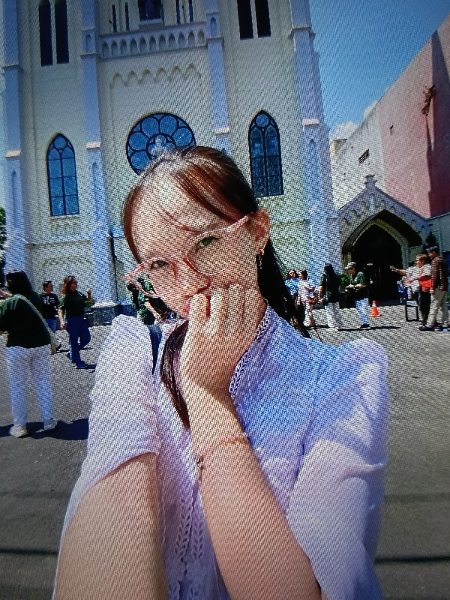 The image is a selfie taken outdoors in front of a white church with a pink building to its right. The church has Gothic architectural elements, including arched windows and a rose window. In the foreground, a young woman with glasses and light brown hair is visible, her face partially obscured by her hand. She is wearing a light purple top with lace detailing. In the background, several people are walking around. The ground is paved. The sky is clear and blue, indicating a sunny day. The scene appears to be in a public space, possibly a plaza or street adjacent to the church. The presence of the church suggests a religious or historical site. The specific location of Raha, Indonesia, is noted as a detail about this scene.