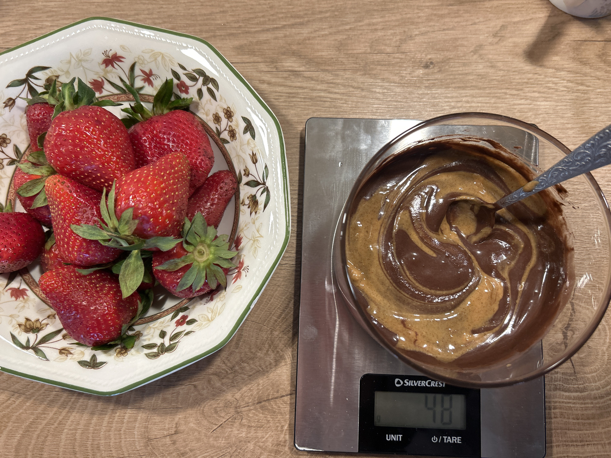 On a wooden surface, a floral patterned bowl of fresh strawberries is positioned next to a glass bowl containing a mixture of chocolate and peanut butter. The glass bowl sits on a digital scale displaying "48". A spoon rests in the chocolate peanut butter mixture. The location in Plovdiv, Bulgaria, cannot be determined from the image.