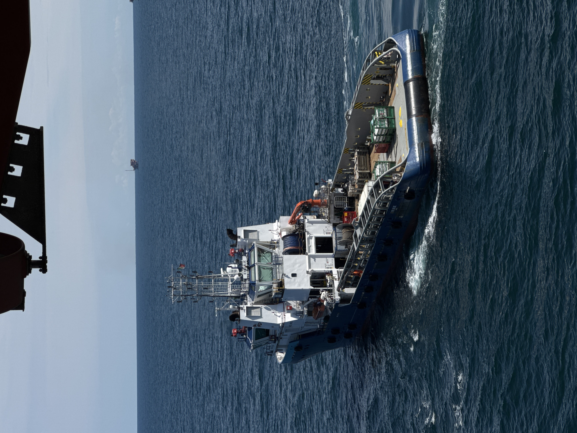 The image shows an aerial view of a blue and white vessel sailing on a calm, deep blue sea under a clear sky. The vessel appears to be a tugboat or a similar working boat, with a white superstructure and a blue hull. There are several crates and equipment visible on the deck of the vessel, along with railings and safety markings. The boat is creating a wake as it moves through the water.
In the distance, a small structure, possibly an oil rig or a platform, is visible on the horizon. The overall scene is one of industrial maritime activity. The lighting suggests it is daytime, with no indication of specific weather conditions beyond the calm sea and clear sky. The image is oriented vertically, with the boat positioned diagonally across the frame, moving from the bottom right towards the top left.
Some text is visible on the side of the vessel, reading "LETO BLUE" and "SUBSEA" with "ITALIA" underneath. There is also some text on the deck near the stern that is partially obscured. No people are clearly visible on the deck. The overall impression is a functional, working vessel engaged in sea operations.