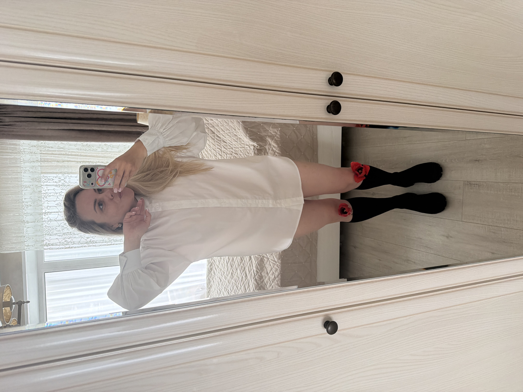 A woman with blonde hair is taking a selfie in a mirror in what appears to be a bedroom. She is wearing a long white shirt and black knee-high socks with red flower decorations. The room is decorated with neutral tones, including a white cabinet, a beige patterned bedspread, and light-colored flooring. Light is entering through a window covered with a lace curtain. The woman is holding a phone with a decorated case. There is no indication in the image that this is in Votkinsk, Russia.