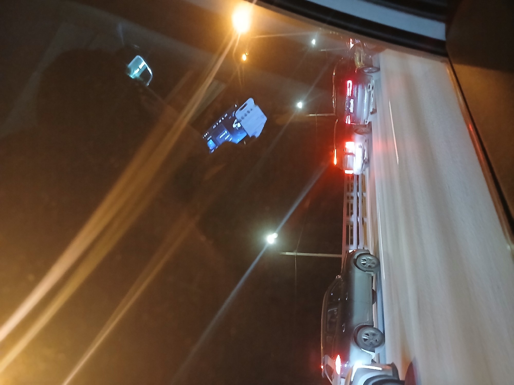 This image depicts a nighttime scene viewed from the interior of a vehicle looking through its front windshield onto a multi-lane roadway, identified as a highway or bridge, located in the city of Unknown, Unknown.
Reflected prominently in the upper center of the windshield is an inverted image of a handgun, featuring a blue upper slide and a white or light-colored lower frame/grip. Additional light streaks and a small, reflective, rectangular object are visible as reflections in the upper portion of the windshield. The dark interior frame of the viewing vehicle is visible along the right and bottom right edges of the image.
Outside the vehicle, several other vehicles are visible on the road, proceeding in the same direction. In the left lane, a partial view of a silver or light-colored SUV is in the immediate foreground, followed by a dark-colored SUV further ahead. In the right lane, multiple vehicles are observed from the rear, including a silver sedan or SUV with illuminated red taillights and a dark-colored SUV with distinctive L-shaped red LED taillights and dual exhaust tips. Overhead streetlights and vehicle illumination provide the primary light sources against the dark environment. White lane markings are visible on the asphalt road surface. No individuals are directly visible in the scene.