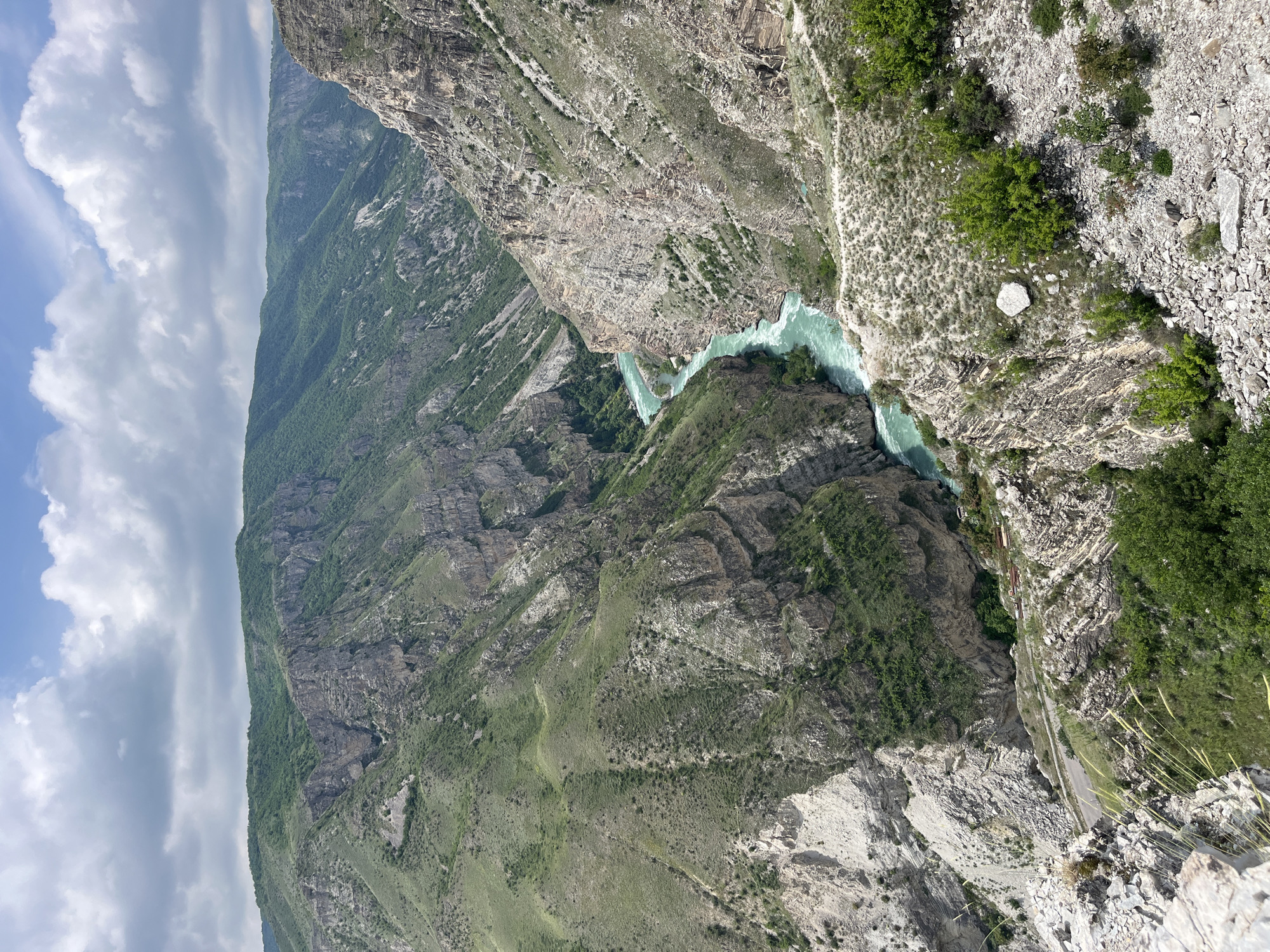 The image depicts a steep canyon landscape with a turquoise river running through it. The canyon walls are a mix of rock formations and green vegetation. In the distance, there are mountains covered in lush greenery under a sky with scattered clouds. Along the cliff edge, a road can be seen. There are no people visible in the image. The location may be near the city of Druzhba, Russia, given the user's request.
