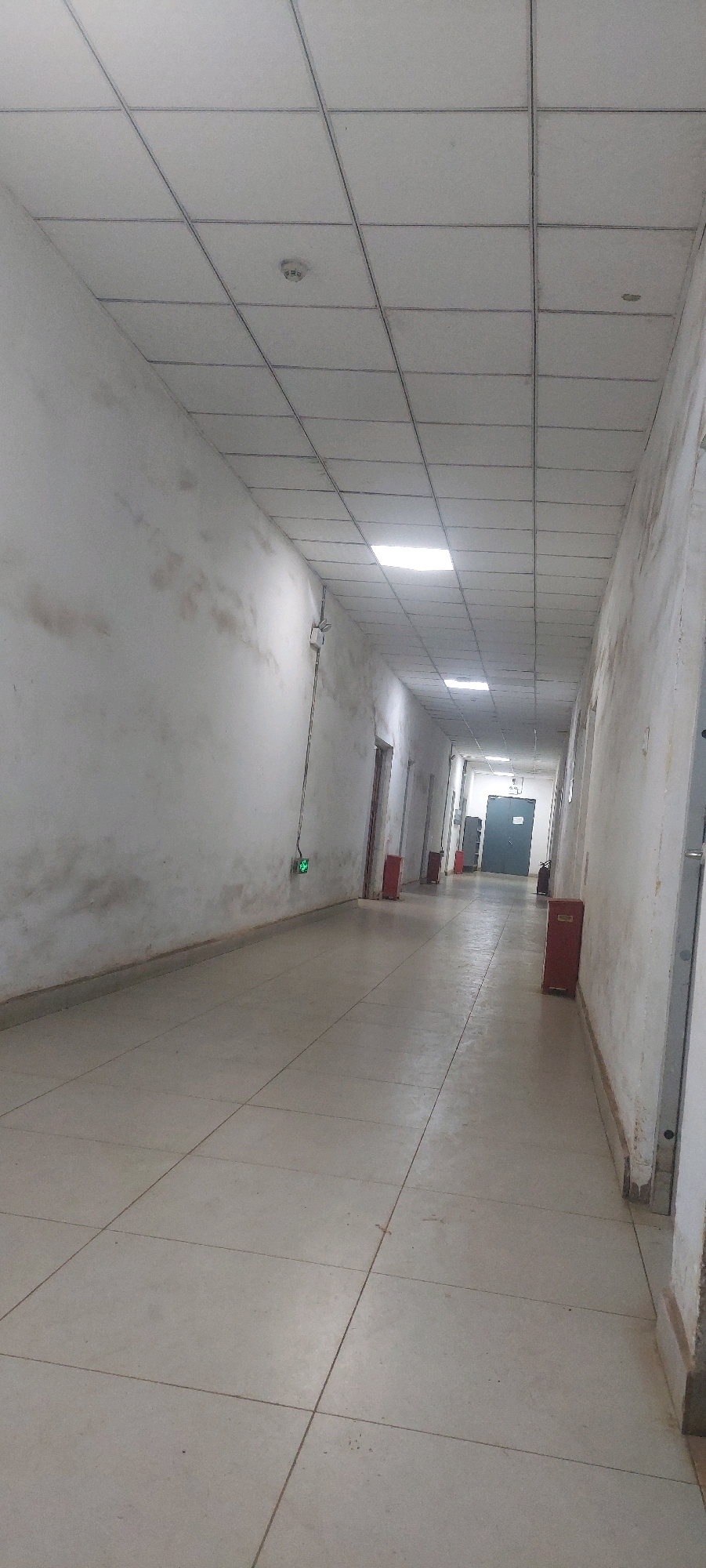 The image depicts a long, indoor hallway with a tiled floor and a dropped ceiling. The walls are a pale, off-white color and appear somewhat dirty or stained in places. Several doors are visible along the right side of the hallway. On the left wall, there is a visible electrical conduit running from the ceiling downwards, terminating in a small, green illuminated sign that reads "EXIT". Further down the hallway on the left wall, a red fire extinguisher is mounted. On the right side of the hallway, closer to the foreground, a red metal bin or cabinet is present. The ceiling is constructed of square tiles with a metal grid structure. Several of these tiles are illuminated by overhead lights, casting a bright glow. There are no people visible in the image. The overall impression is that of a utilitarian, institutional hallway.