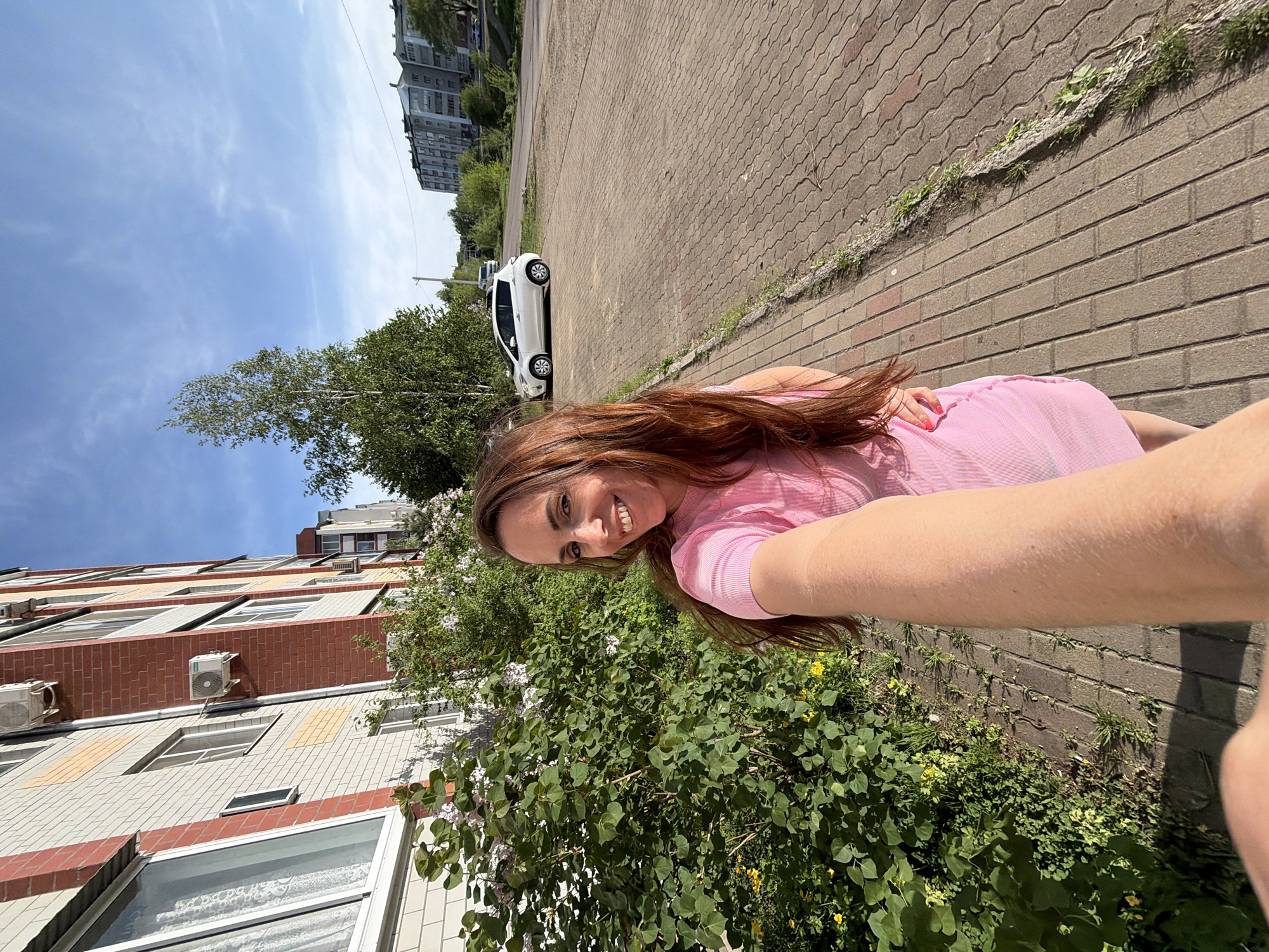 A woman is taking a selfie in Birobidzhan, Russia. She is wearing a pink dress. Behind her is a brick sidewalk and a street with a white car. A building with air conditioning units is to the left, and trees are in the foreground. The sky is partly cloudy.