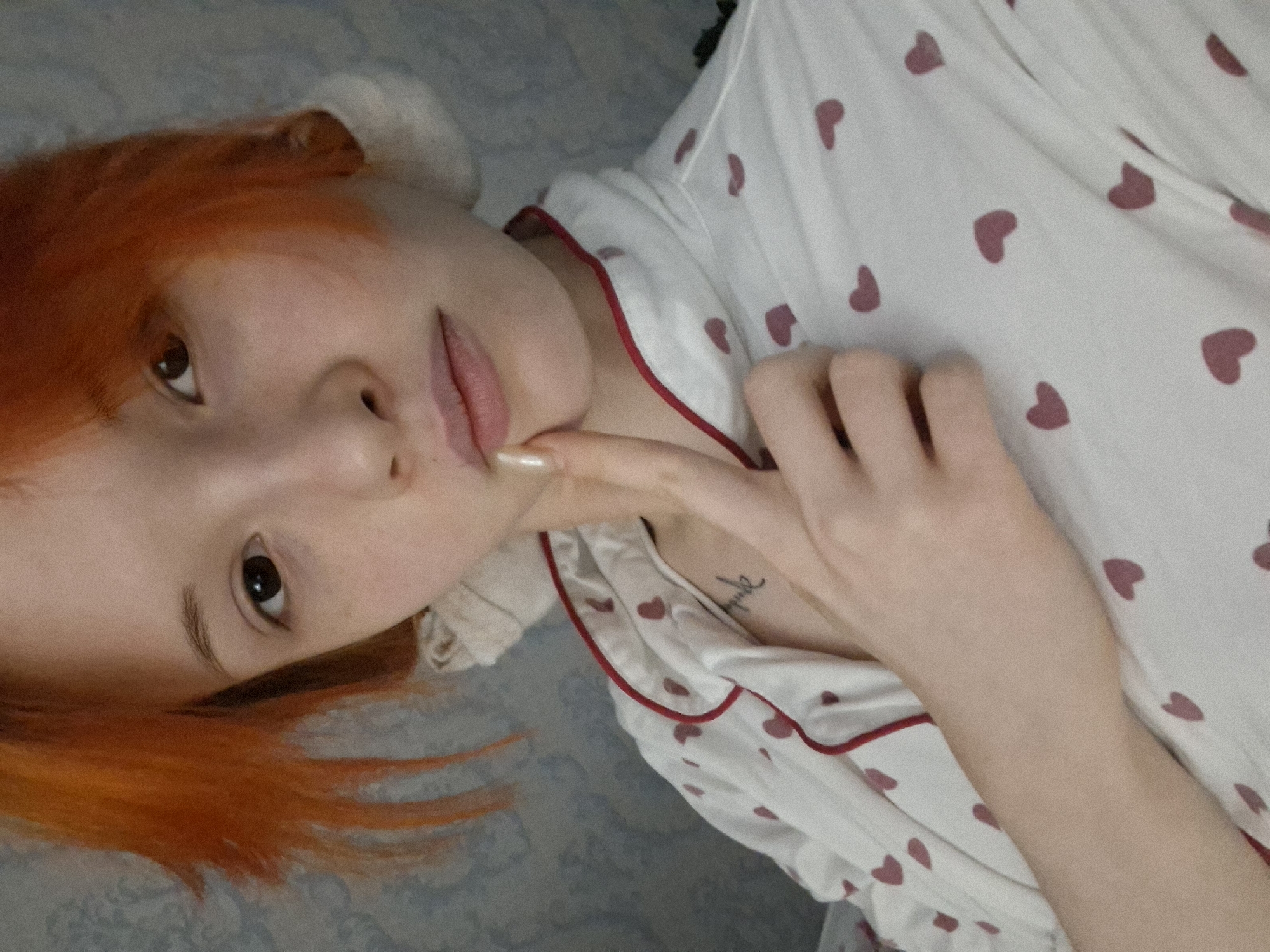 A fair-skinned individual with red hair is lying down on a blue and white patterned surface. They are wearing a white pajama top with red heart prints and red piping. Their right hand is raised towards their face, with their index finger touching their lips. A small tattoo is visible on their chest. The background appears to be a bed. There is no verifiable evidence to definitively place the location in Chaikovsky, Russia.