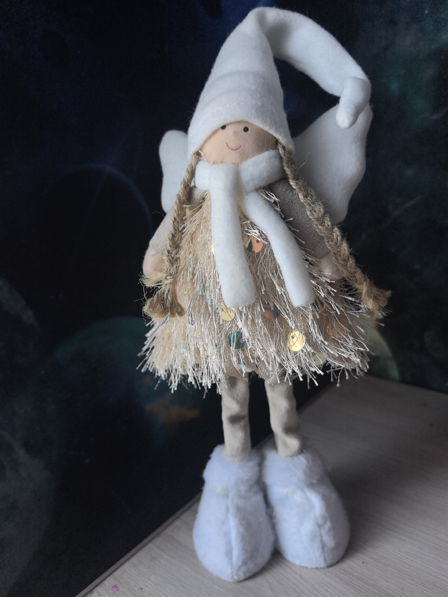 A fabric doll, styled as an angel or elf, stands upright in a static pose. The doll features a light beige, smooth fabric face with two embroidered black dots representing eyes and a red curved line for a smile. It wears a white pointed hat, made of a soft material like felt, which bends to the right. A white scarf of similar material is wrapped around its neck. Two braided pigtails, constructed from a rough, light brown cord, extend from either side of its head. The doll's body is covered by a skirt-like garment composed of numerous thin, light-colored, shaggy fibers, interspersed with small, reflective multi-colored sequins. White, felt-like wings are attached to its upper back, partially visible behind its shoulders. Its legs are thin and light beige, leading to large, white, soft boot-shaped feet. The doll is positioned on a light-colored surface exhibiting a faint horizontal wood grain pattern. The background is dark, out-of-focus, and devoid of discernible features. The image provides no visual information to determine the location within the city of Sarapul, Russia.