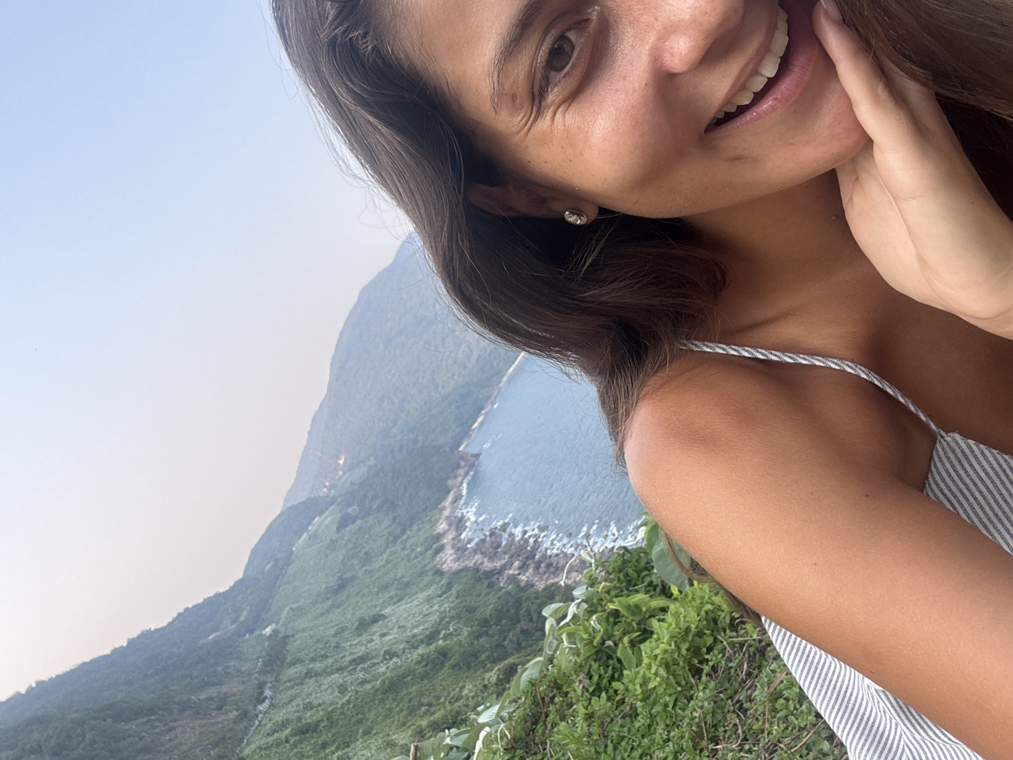 A selfie-style portrait of a smiling woman is framed against a backdrop of a lush mountain landscape meeting a coastal area. The woman has brown hair, a light complexion, and is wearing a striped sleeveless top. Her hand is raised to her face, and she is looking directly at the camera.
The background reveals a steep, verdant mountain range sloping down to a rocky coastline. A body of water, likely the ocean, is visible, and the sky is a pale blue. The photograph appears to have been taken at a high vantage point.
This is a location in the city of Đà Nẵng, Vietnam.