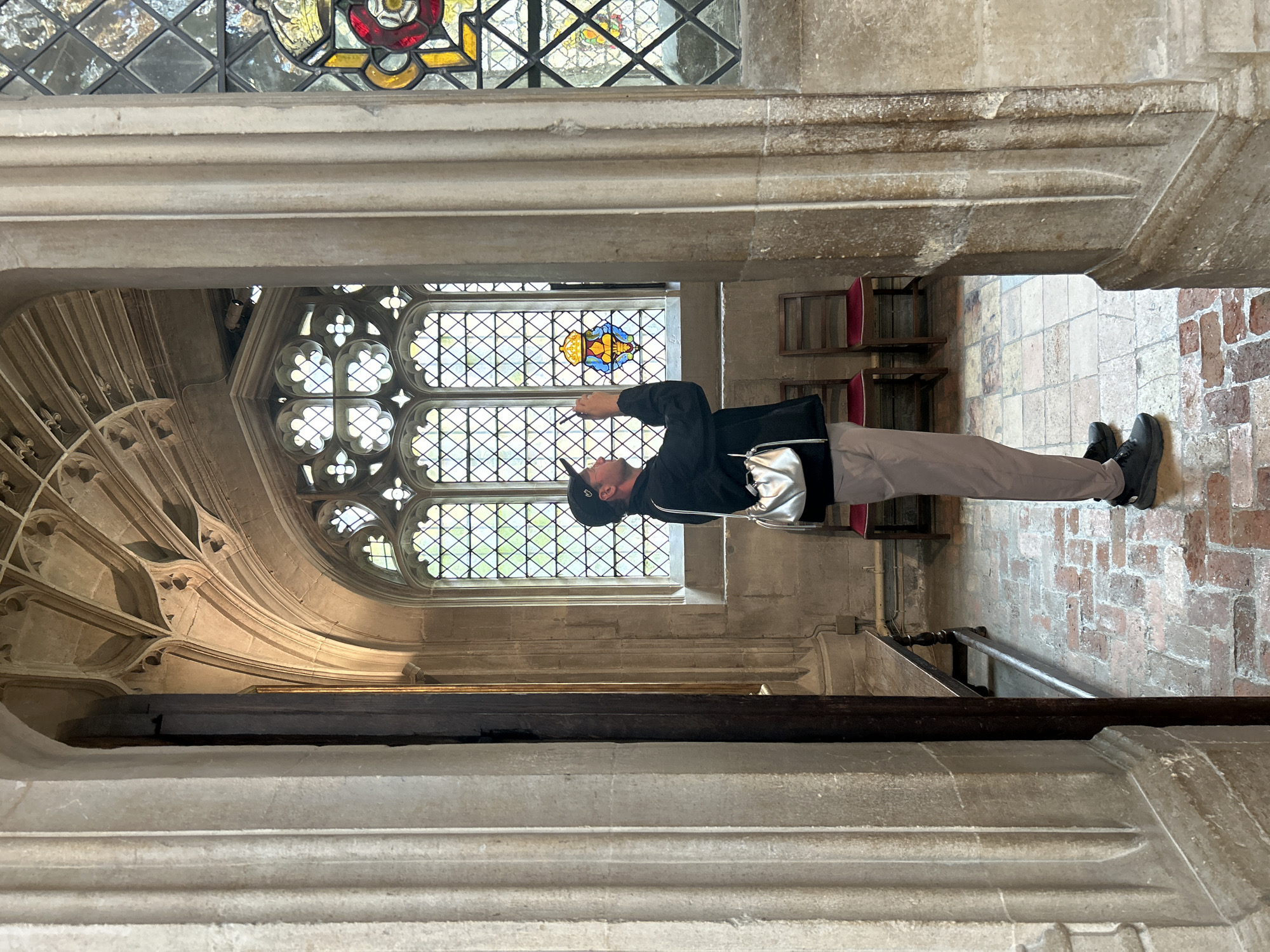 A person is standing in an arched entryway, looking into a room with stained glass windows. The person is wearing a black baseball cap, black jacket, and light-colored pants. They are holding a phone up to take a picture of the stained glass. In the background, there are two wooden chairs with red seats, a wooden bench, and a brick floor. The ceiling has intricate stone carvings. This scene appears to be inside a historic building, potentially King's College Chapel in Cambridge, United Kingdom, given the architectural style.
