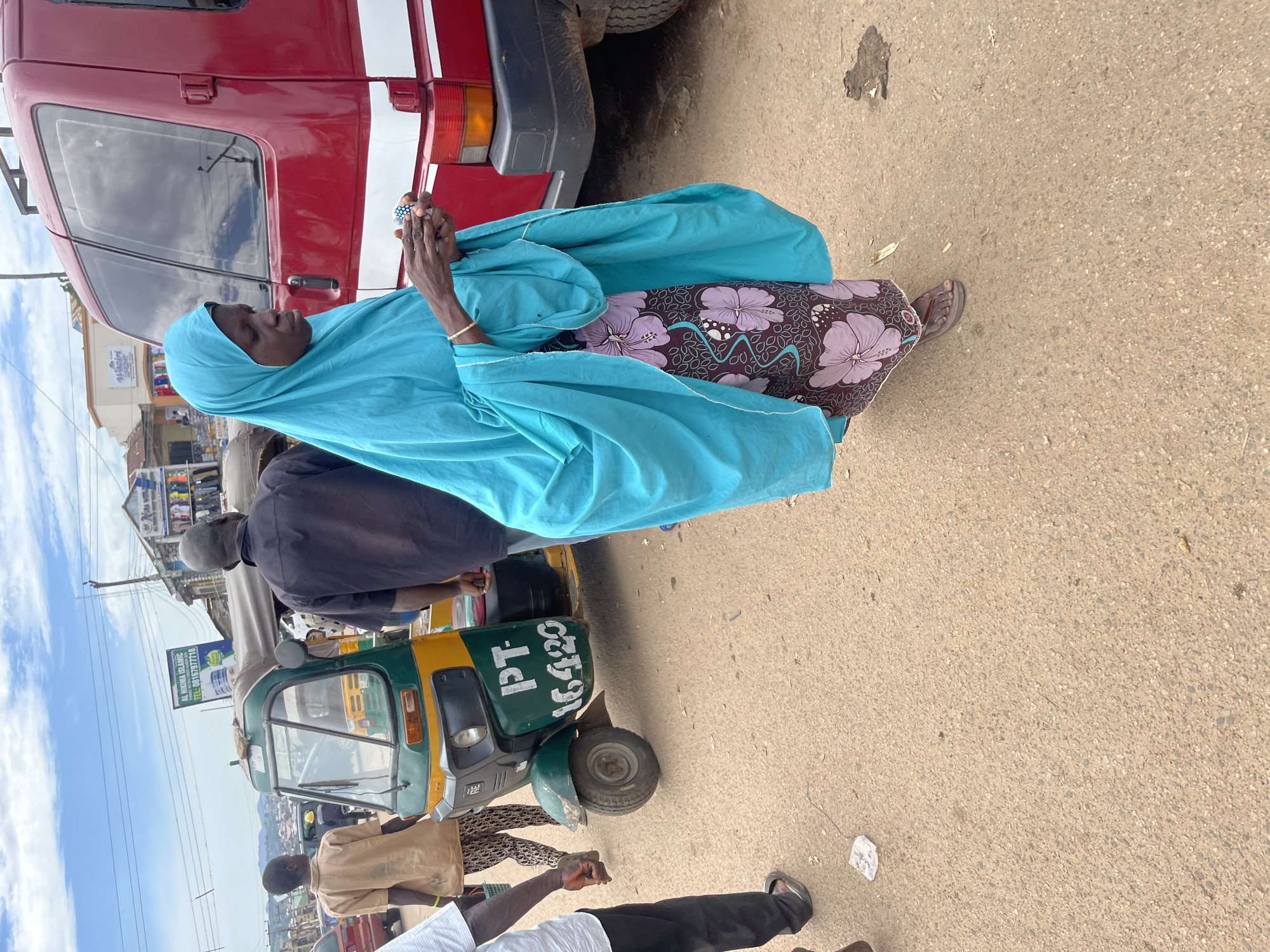 A medium shot shows a woman in a bright turquoise hijab and a patterned skirt walking on a paved street in Jos, Nigeria. She is looking down at something in her hands, which appear to be small. The woman is walking alongside a red van. In the background, a green and yellow auto-rickshaw is parked. Further behind the auto-rickshaw, a sign for "AL HIKIM ISLAMIC" is visible, along with several buildings and a cloudy sky. The street is mostly clear, with a few small pieces of debris scattered on the ground. The overall scene depicts a street environment in a Nigerian city.
