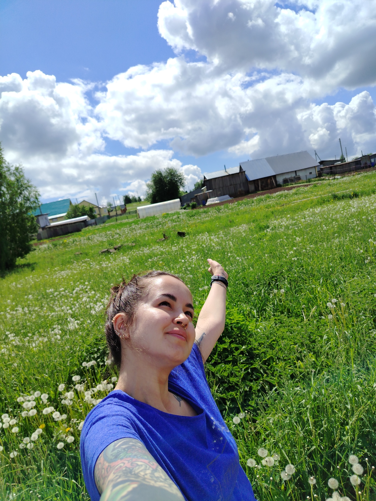 A young woman stands in a grassy field under a partially cloudy sky, appearing to take a selfie with an outstretched arm. She has her hair pulled back, wears a blue shirt, and has visible tattoos on her arm. The field is densely covered in grass, with scattered dandelion seed heads. In the background are several wooden buildings typical of rural architecture. The location appears to be the city of Гожан, Russia, based on the architecture and general scenery. The overall scene is captured in bright daylight.