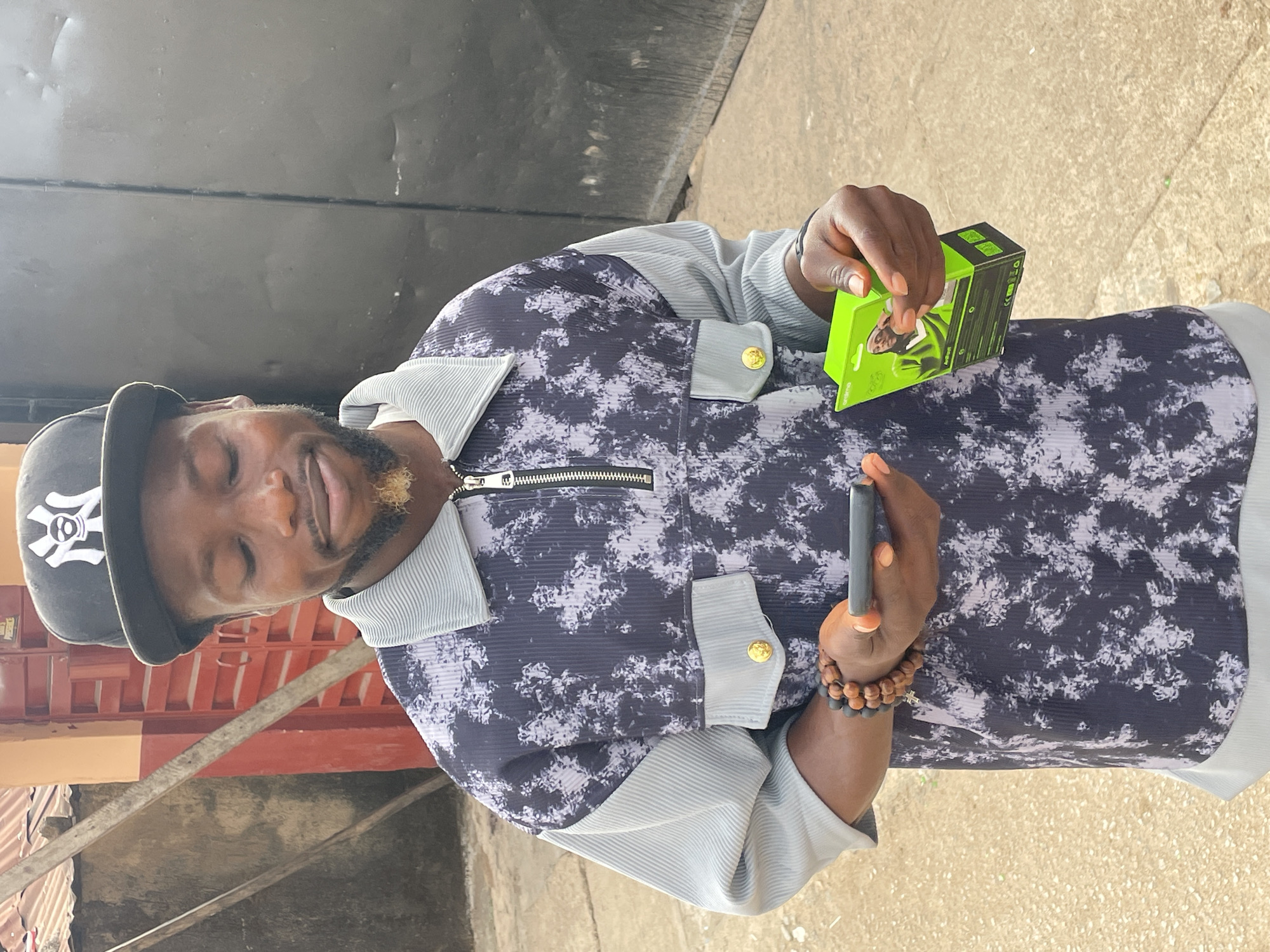 A man stands in what appears to be an outdoor setting in Jos, Nigeria. He is wearing a black baseball cap with a white "NY" logo, a patterned shirt with ribbed sleeves and gold buttons, and a beaded bracelet. He is holding a gray vape pen in his left hand and a green box, possibly containing the vape, in his right hand. The background includes a dark metal door and a red-painted structure. The ground appears to be paved.