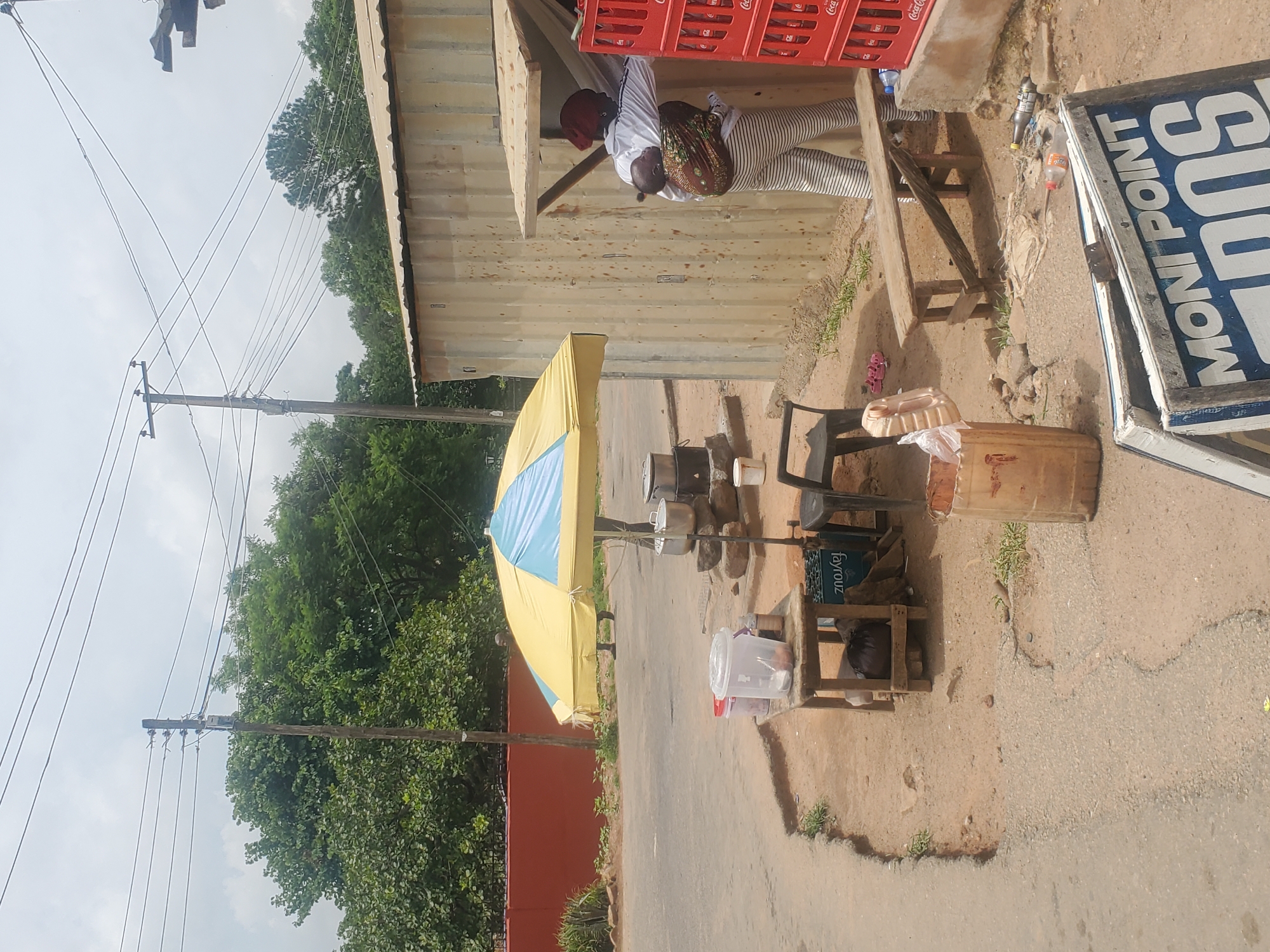 An outdoor scene in Jos, Nigeria depicts a small business operation. A woman carrying a baby on her back stands near a corrugated metal kiosk, wearing a red headscarf, a white jacket, and striped trousers. She appears to be interacting with the interior of the kiosk. In front of the kiosk, a yellow and blue umbrella shades a table laden with containers and what appear to be cooking pots. A wooden bench is situated to the right of the kiosk, and stacks of Coca-Cola bottles are visible. A wooden sign with "MONI POINT" is in the foreground. The ground is dirt and uneven, with some debris scattered around. Lush green trees and telephone poles are visible in the background, suggesting an urban or semi-urban environment.