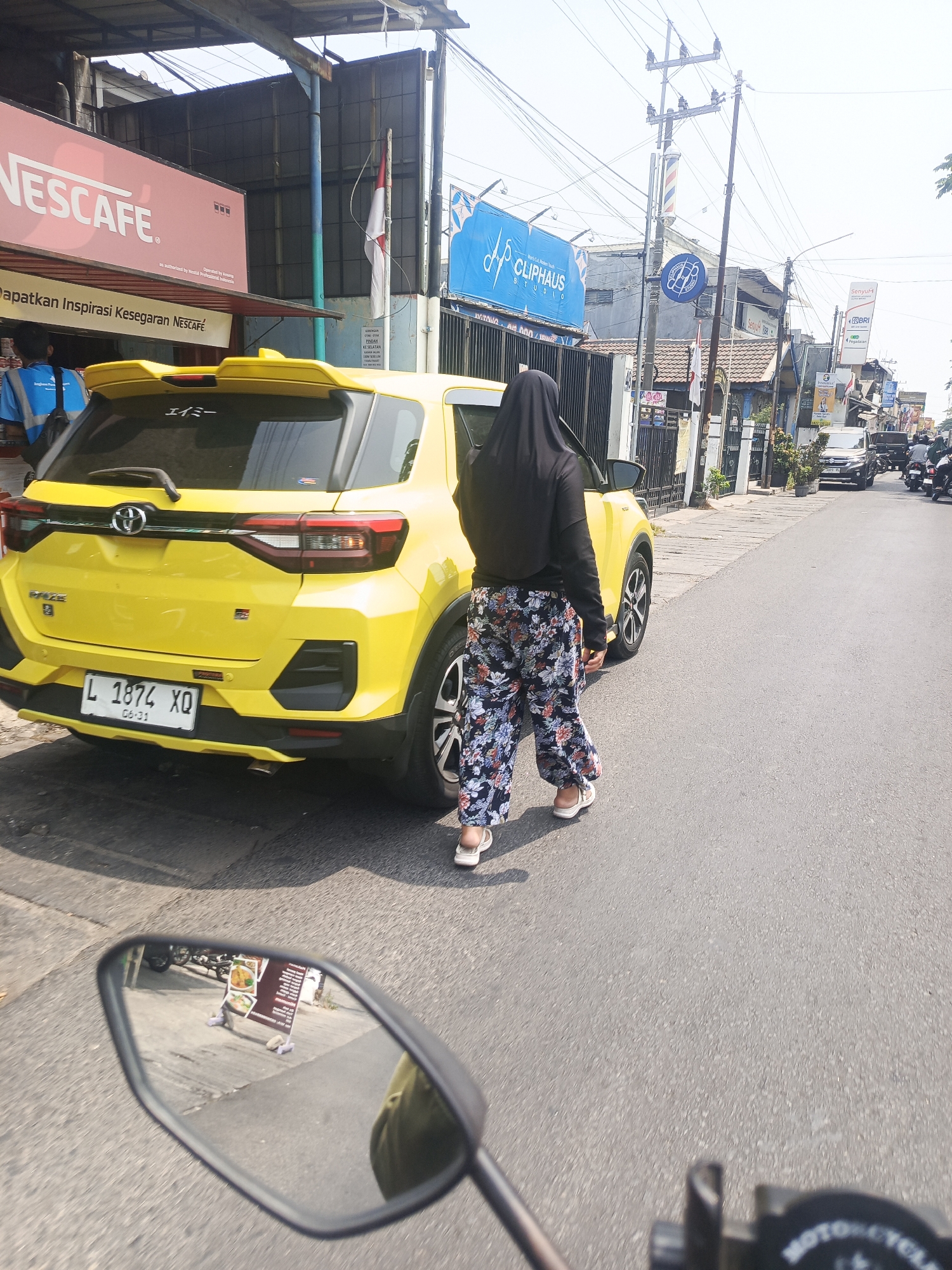 The image depicts a street scene in Sedati Gede, Indonesia. In the foreground, a yellow car, identified by its license plate "L 1874 XQ", is parked on the side of the road. A person wearing a black hijab and floral patterned pants is walking away from the car towards the right side of the frame. The car's rear is visible, showing taillights and a spoiler. The surrounding environment includes buildings with various signs, including "NESCAFE" and "CLIPHAUS STUDIO". Utility poles and overhead wires are prominent in the background. The road appears to be asphalt. A motorcycle mirror in the lower left corner reflects a sign with images of food and text. Other vehicles, including motorcycles and cars, are present further down the street, suggesting moderate traffic. The sky is bright, indicating daytime.