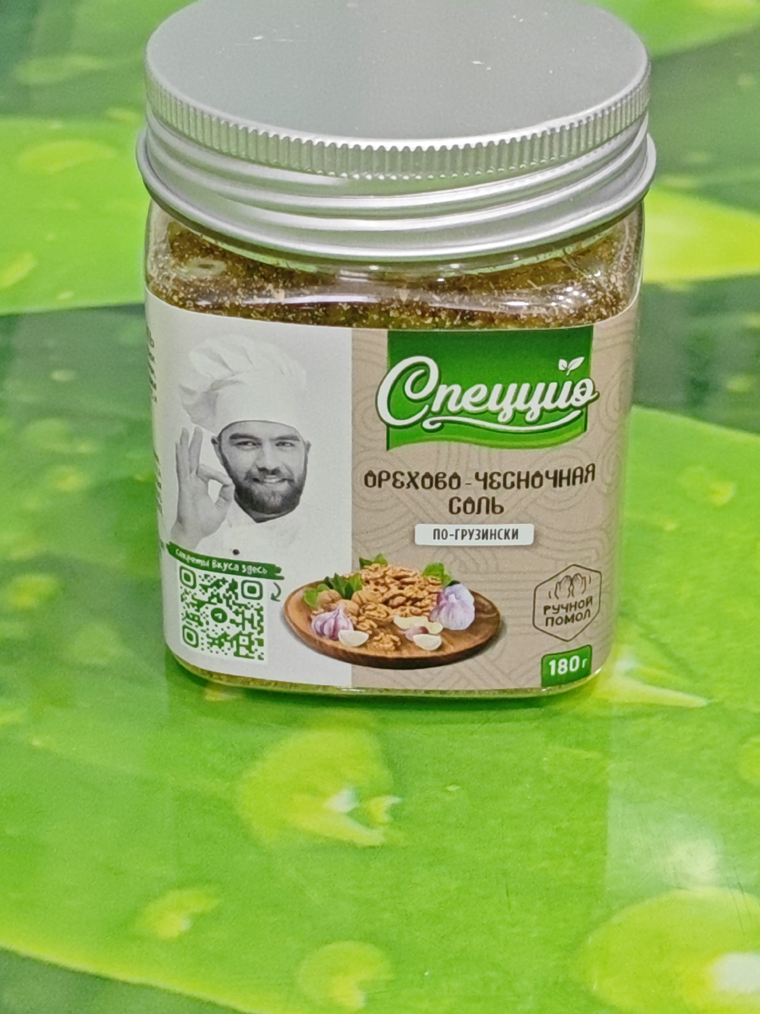 A close-up, slightly angled shot shows a glass jar of seasoning with a silver lid, placed on a bright green, textured surface. The jar's label features a chef's illustration, a QR code, and text in Russian. The text on the label reads "Специй", "ОРЕХОВО-ЧЕСНОЧНАЯ СОЛЬ", "ПО-ГРУЗИНСКИ", "РУЧНОЙ ПОМОЛ", and "180г". The seasoning inside the jar appears granular and speckled. The background is a blurred, vibrant green, creating a sense of being outdoors or on a patterned surface. There are no discernible people or actions captured within the frame. The image does not provide any information about the city of Polyarny, Russia.