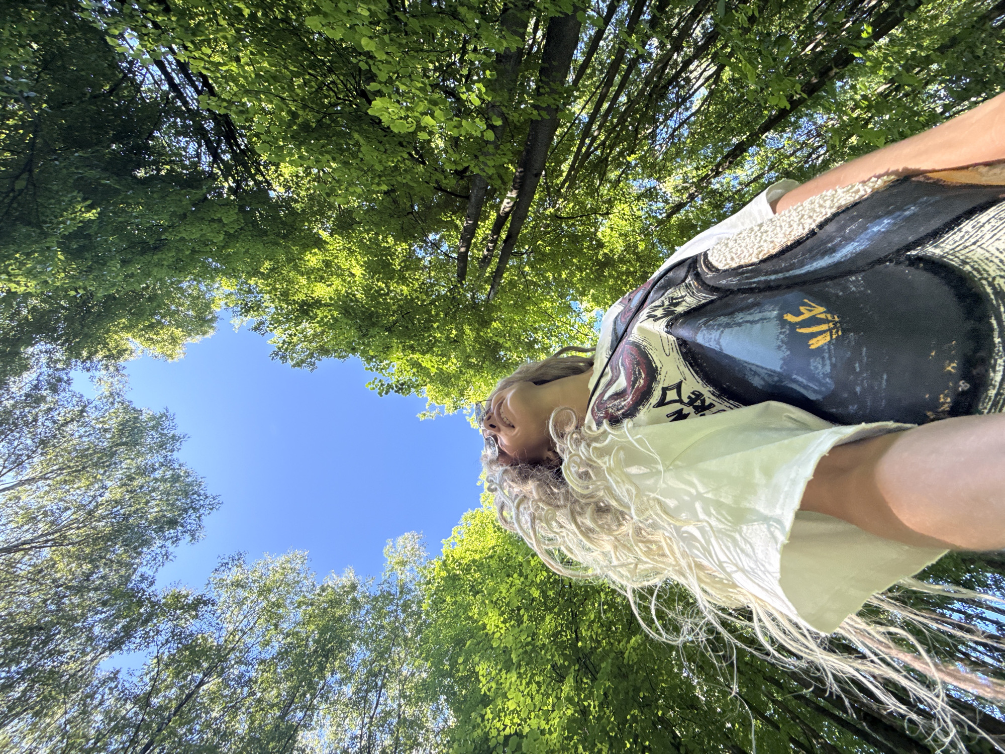 A low-angle outdoor photograph depicts a person, likely female, with long, light-colored dreadlocks or very curly hair, looking upwards into a clear blue sky in Rusanovo, Russia. The subject wears sunglasses positioned on her head, a white short-sleeved t-shirt, and a dark-colored, patterned sleeveless vest or overlay. Dense, vibrant green tree foliage, illuminated by direct sunlight, surrounds the central patch of clear blue sky. The perspective is from beneath the tree canopy, looking up at the subject and the sky through the leaves and tree trunks.