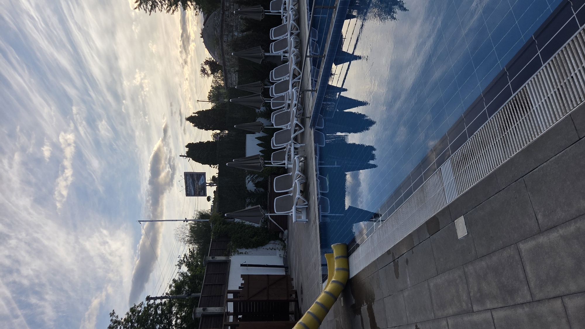 This image depicts an outdoor swimming pool area at dusk or dawn. The pool is filled with blue water and reflects the sky, which features wispy, streaky clouds. Several empty lounge chairs are arranged in rows next to the pool, each with a closed umbrella. A yellow water slide is visible on the left side of the image, extending into the pool area. The poolside is paved with dark grey tiles. In the background, a fence encloses the area, and beyond that, there are trees, utility poles, and a billboard advertising "Pulse Therme" with an image of a pool. The overall environment appears to be a recreational facility, possibly a hotel or public spa, in an urban or suburban setting. There are no people present in the scene.