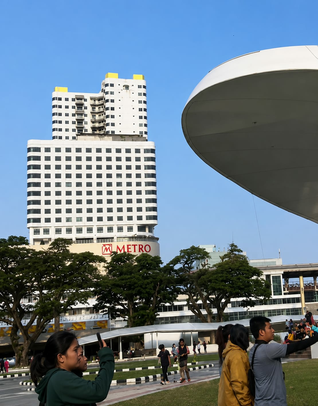 In a wide-angle shot of a city park, the Metro department store's building is prominent with its large red logo against a white facade. The building has a distinctive stepped design with yellow accents on the upper levels. To the right, a large, curved white structure partially obscures the sky, suggesting a modern architectural feature. A clear blue sky serves as the backdrop. In the foreground, several people are seen walking and taking pictures. One woman in a dark green hoodie is focused on her phone, possibly taking a selfie or recording. Other individuals are also engaged in various activities, with some appearing to be tourists. The park area features a paved walkway with black and white borders, and lush green grass. Mature trees with dense foliage are scattered throughout the scene, adding a natural element to the urban landscape. The scene appears to be captured during daylight hours, with soft lighting indicating it might be late afternoon. The overall impression is of a vibrant public space where modern architecture and nature coexist, and people are enjoying leisure time. This location is likely the Lapo Tonda (Tonda Fountain) area within the city of Medan, Indonesia, known for its modern amenities and recreational spaces.