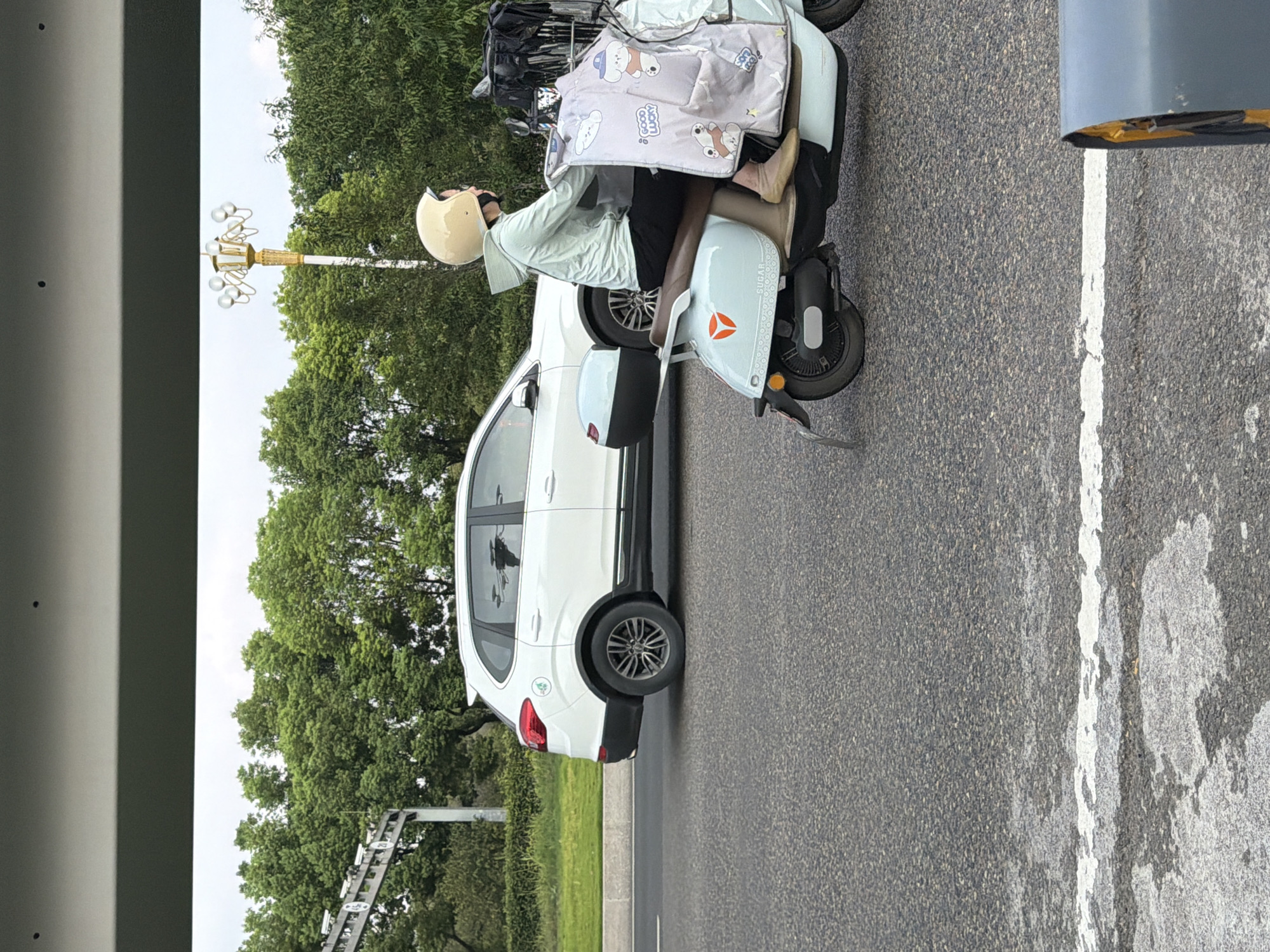 A white SUV is driving on a paved road in Jiangjin, China, next to a pale blue electric scooter carrying a person wearing a light blue hoodie and a cream-colored helmet. The scooter has a decorative blanket with cartoon dog illustrations and the words "GOOD LUCKY" printed on it draped over the front. The background features lush green trees and a cloudy sky. The road also has white lane markings.