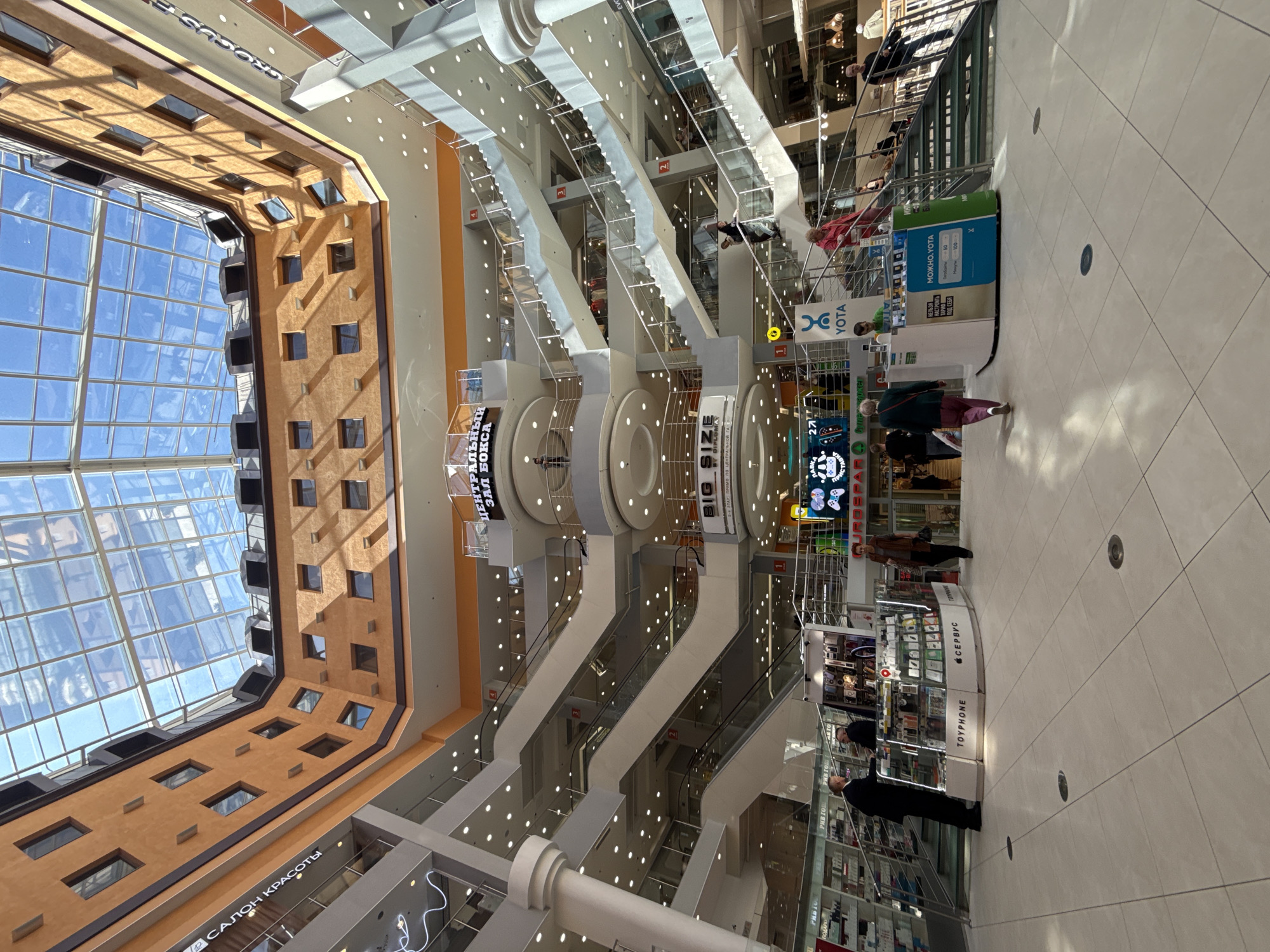 This image shows the interior of a multi-story shopping center with a large glass atrium ceiling. The architecture is modern, with orange-colored walls and multiple levels of escalators, staircases, and walkways. Various shops and kiosks are visible on different floors, displaying a variety of goods. People are seen walking around, using escalators, and standing at service counters. One kiosk in the foreground displays "TOYPHONE" and "СЕРВИС" branding, suggesting it sells or repairs phones. Another kiosk features the "YOTA" logo. The overall impression is a busy and well-lit commercial space. The sign in the background reads "ЦЕНТРАЛЬНЫЙ ЗАЛ ВОКСА" and "BIG SIZE," indicating a central hall, possibly related to a cinema or a large clothing store. The presence of Cyrillic text on signs, such as "САЛОН КРАСОТЫ" (Beauty Salon) and "РЫВ ГОТО" (likely a brand or store name), strongly suggests the location is in Russia. Given the architectural style and common shopping center designs in Russia, and considering the context of a multi-level atrium and specific brand signage, this scene is highly likely to be the **Central Children's Store on Lubyanka** in Moscow, or a similar large shopping complex in Russia, rather than Saint Petersburg. However, without definitive explicit visual cues linking it to Saint Petersburg, a precise identification to that city is not possible.