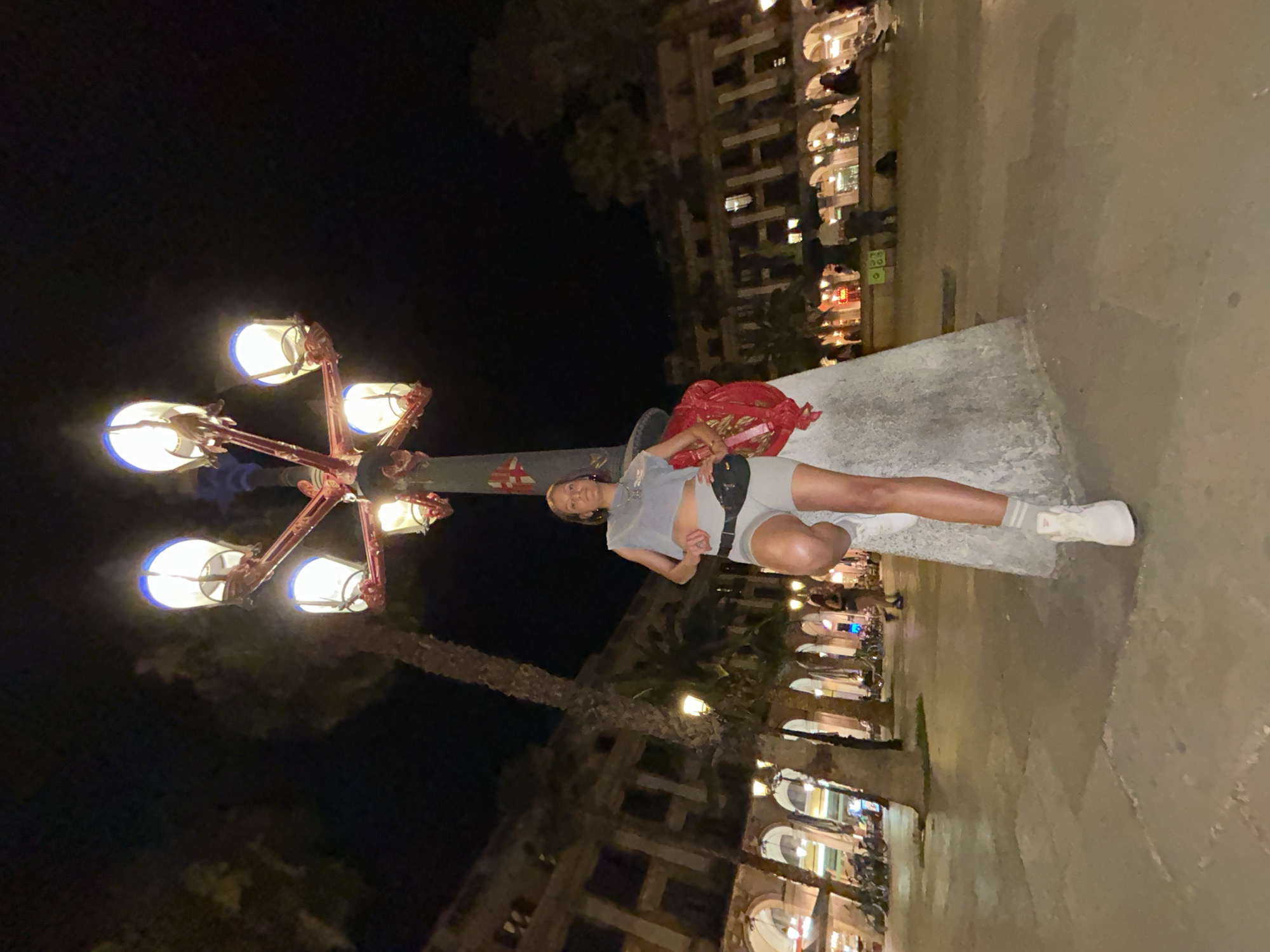 A woman is posing at night in front of a large, ornate lamppost in a public square in Barcelona, Spain. She is wearing a grey crop top, grey shorts, a black fanny pack, white sneakers, and striped socks. The lamppost has multiple lights and is adorned with decorative elements. In the background, there are palm trees and buildings with arched walkways, and several people can be seen walking and sitting in the square. The overall scene suggests a lively urban environment at night.