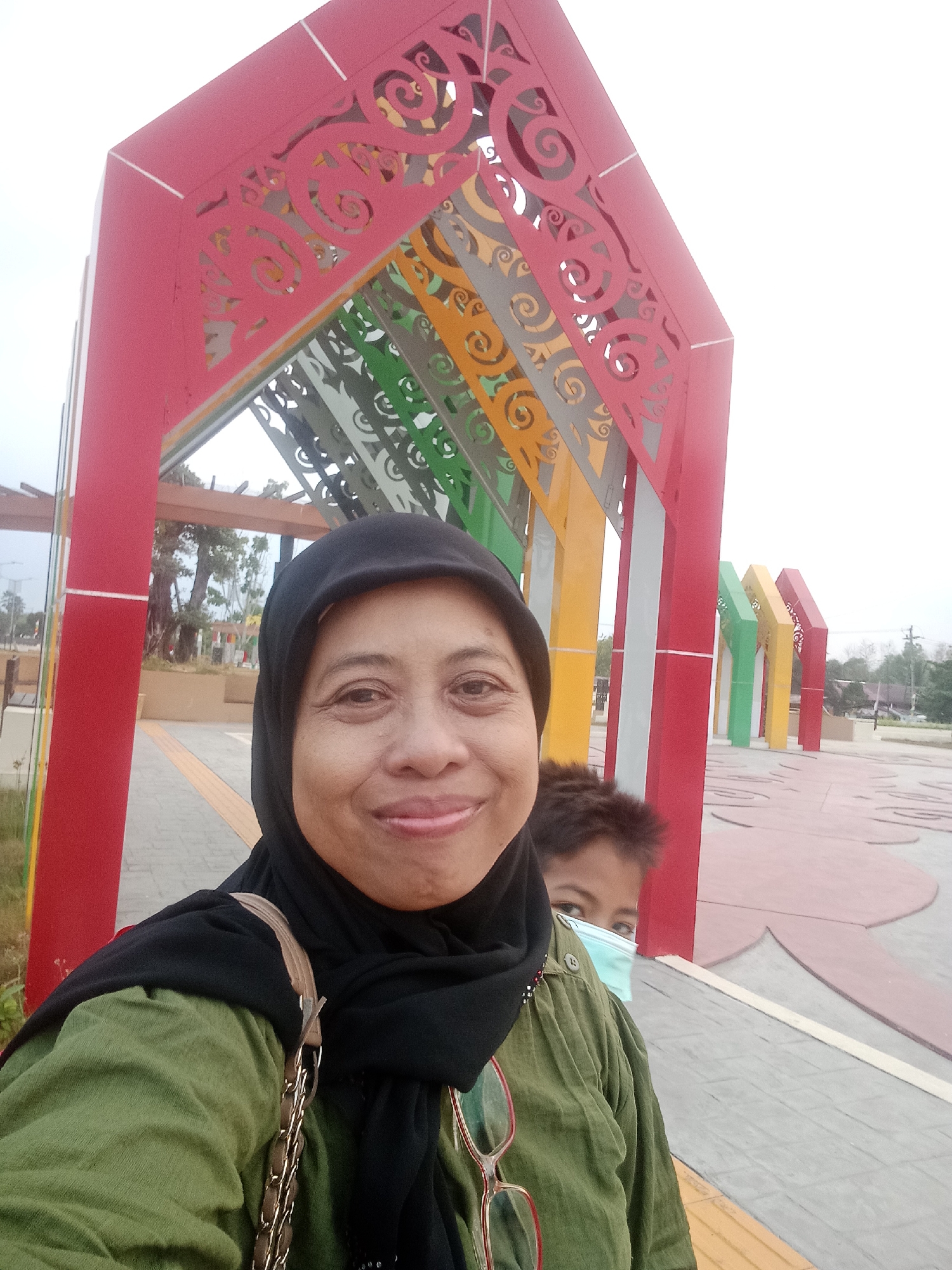 A medium shot selfie shows two individuals in an outdoor urban setting with a colorful architectural structure. In the foreground, a woman wearing a black hijab and a green long-sleeved shirt smiles at the camera. She has glasses perched on her shirt. To her right and slightly behind her, a young boy with dark hair is partially visible, wearing a light blue surgical mask. They are standing on a paved walkway with a textured pattern. Behind them, a large, abstract structure composed of red, green, yellow, and grey arched elements forms a canopy. The background reveals trees and a wide-open area with a decorative pattern on the ground. The overall scene suggests a public park or recreational space in Palangka Raya, Indonesia, possibly along the Sabangau River due to the distinctive architectural style often seen in the city's public spaces.