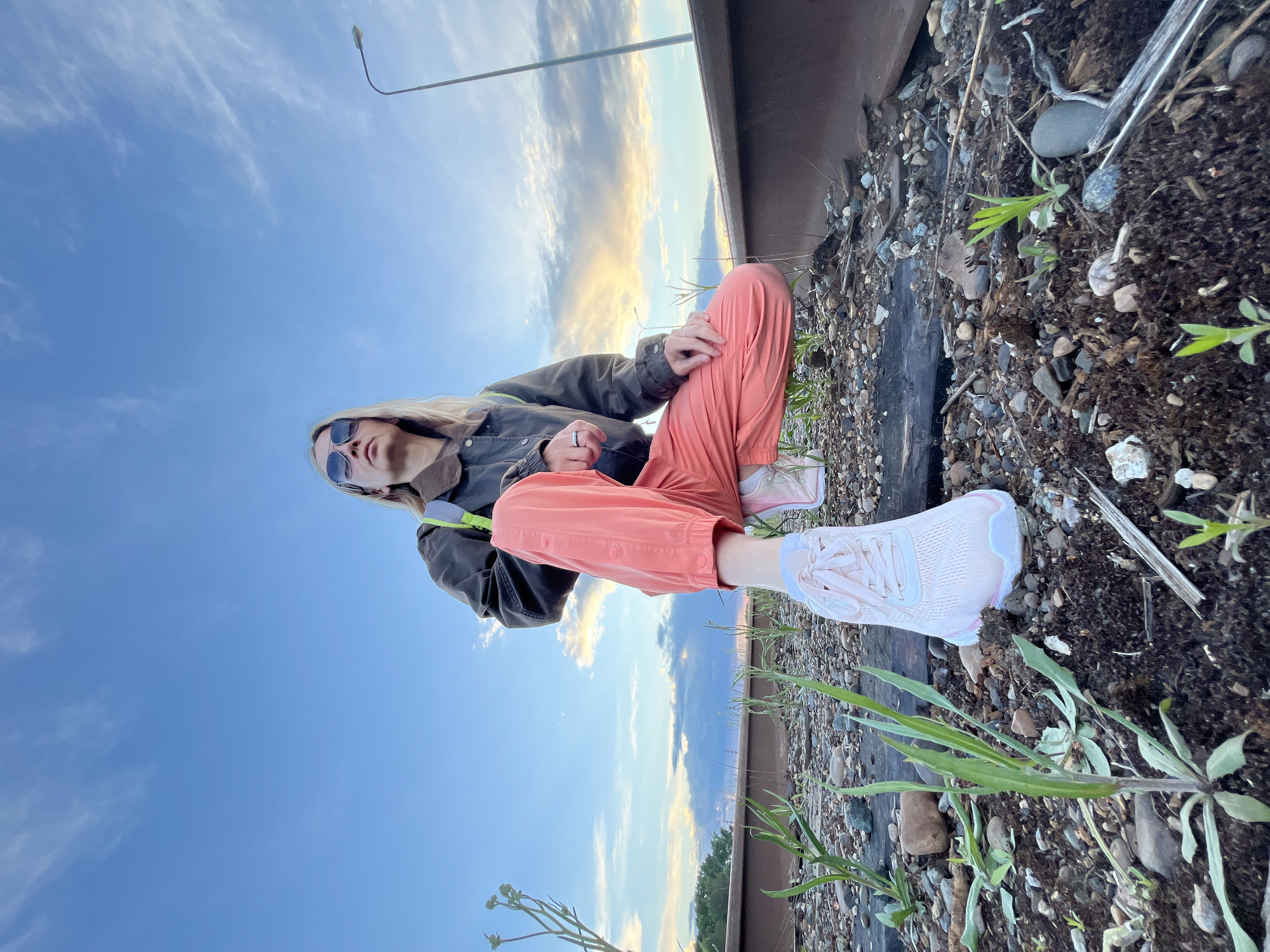 A young woman sits cross-legged on the ground near a set of railroad tracks, against a backdrop of sky and a light pole. She is wearing a dark jacket, coral-colored pants, and light-colored sneakers. Sunglasses obscure her eyes. The ground is a mix of dirt, gravel, and vegetation, including green plants. The sky shows hues of blue and some sunset colors. The photo appears to have been taken at an angle. The specific location in the city of Chaikovsky, Russia is indistinguishable without further information.