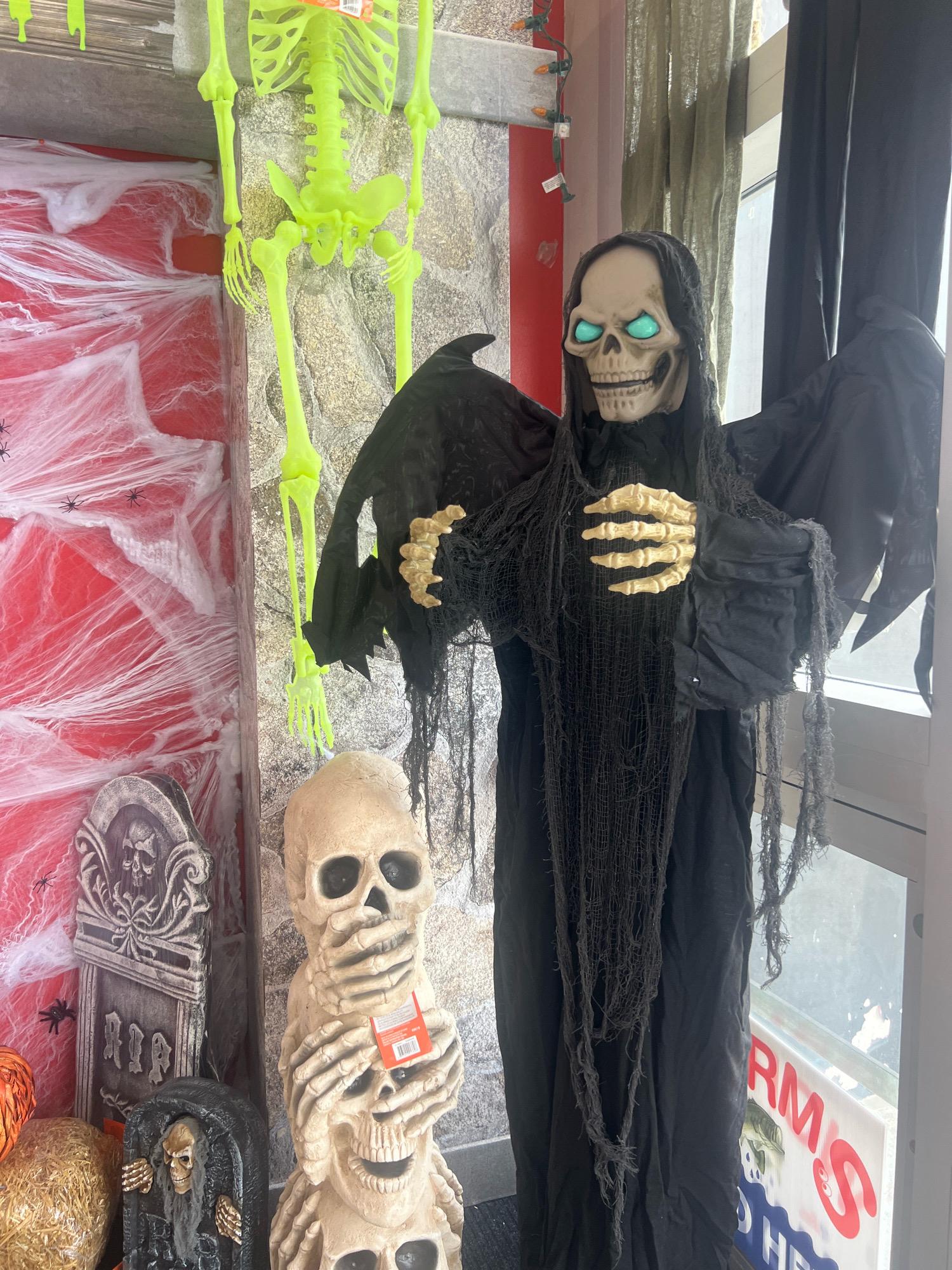 The image shows a Halloween display outside a store, possibly in Toronto, Canada. In the foreground, a grim reaper decoration stands with its arms outstretched. It is wearing a black hooded robe and has a skull for a head with glowing blue eyes. To the left of the grim reaper, a stack of three skulls is visible, with the top two covering their eyes and mouth. Behind these figures, a tombstone decoration and a skeleton hang from the ceiling. The backdrop includes a red wall adorned with white cobwebs and some small spider decorations. A large window with a white sign featuring red lettering is visible in the background. The overall scene is designed to evoke a spooky Halloween atmosphere.