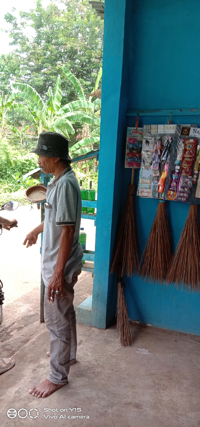 An older man with a dark hat, light gray shirt, and gray pants stands barefoot outside a blue structure. He holds a cigarette in his hand. A motorcycle sits to his left. Behind him are trees and greenery. On the blue structure, hanging are brooms and various packaged items. Bottles filled with green liquid are seen on a shelf. "Shot on Y15 Vivo Al camera" appears in the lower left corner.