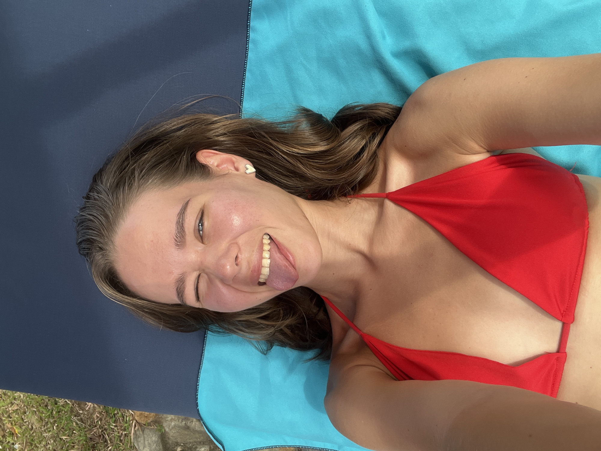 A young woman lies on a two-toned blue and black towel, possibly on a beach. She wears a red bikini top. Her hair is long and brown, styled in loose waves. She is smiling and sticking out her tongue. She has a heart-shaped earring in her left ear. The bottom left corner of the image shows grass and a rock, suggesting an outdoor location.
The location details provided are “Nam Nha Trang Ward, Vietnam.” While the image suggests an outdoor setting, it is difficult to pinpoint the exact location within Nam Nha Trang Ward.