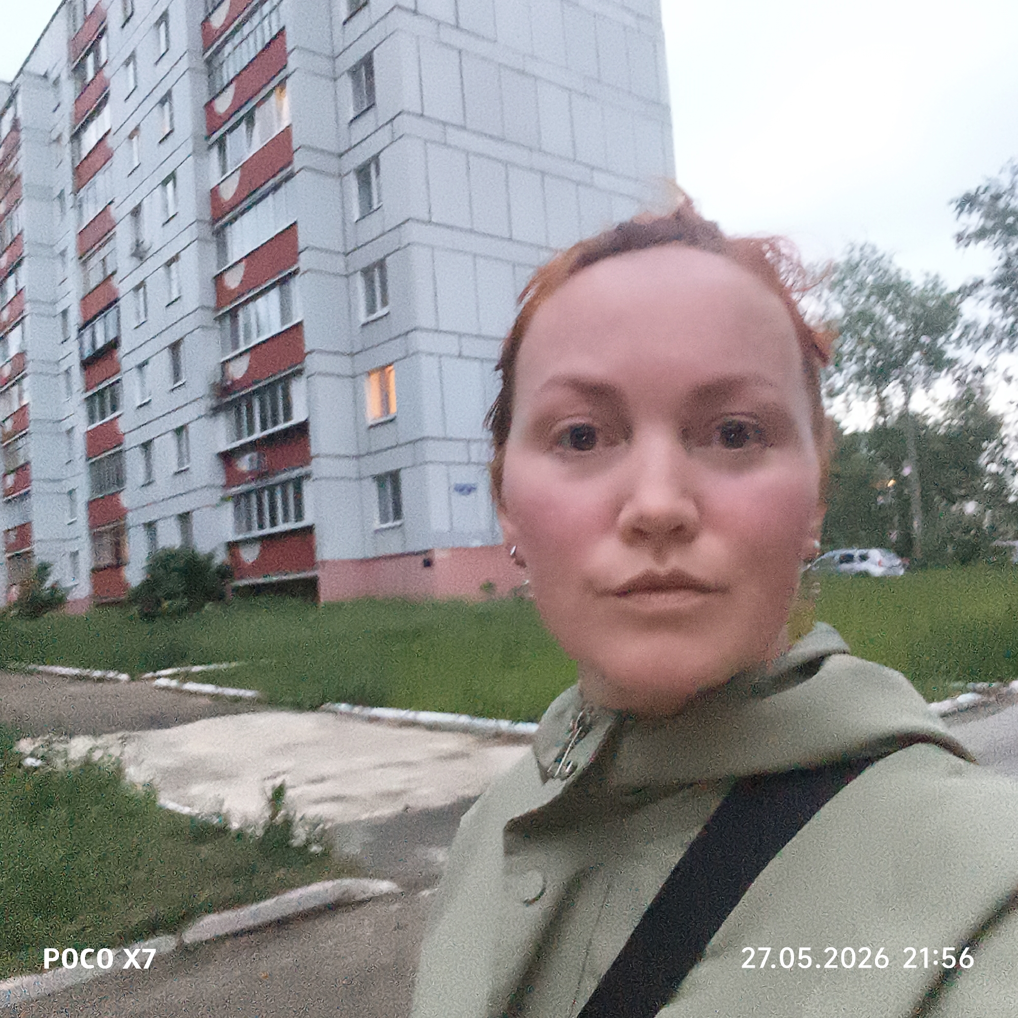 The image shows a selfie of a fair-skinned person with reddish hair. The person is wearing a light green jacket with a dark strap across the chest. Behind the person is a large, multi-story apartment building with a light gray facade and red balcony accents. A lit window is visible on one of the floors. In the background, there is green grass, trees, and a car. The date and time "27.05.2026 21:56" and the device "POCO X7" are displayed in the corners of the image. The setting appears to be an urban residential area. The location is likely in Chaikovsky, Russia.