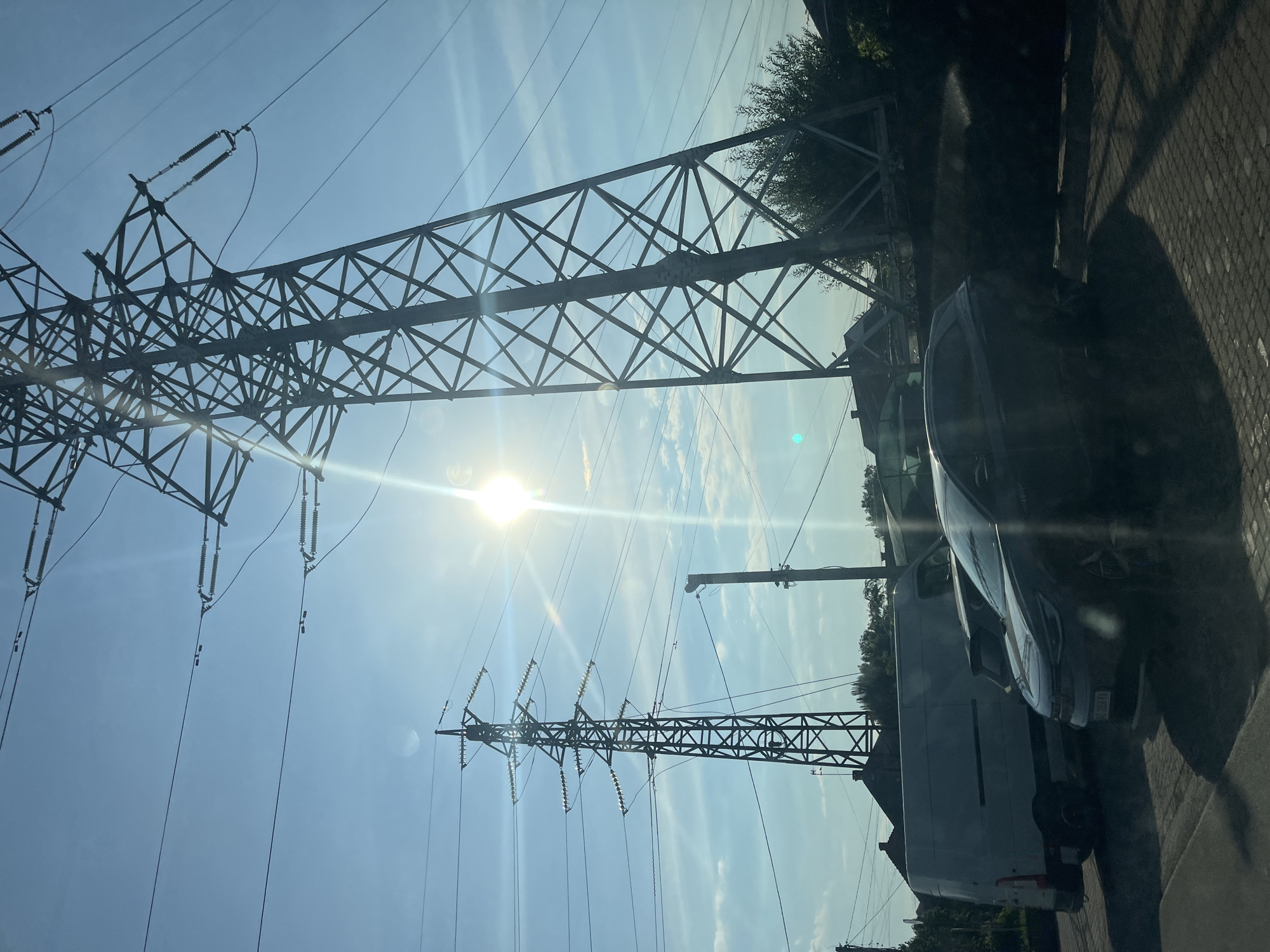 In Storozhnytsia, Ukraine, a large electricity pylon stands prominently against a clear blue sky with a bright sun that causes lens flare. Power lines extend from the pylon across the frame. In the foreground, a dark-colored car is parked on a cobblestone surface, with a white van partially visible behind it. The scene includes scattered trees and rooftops in the background.
