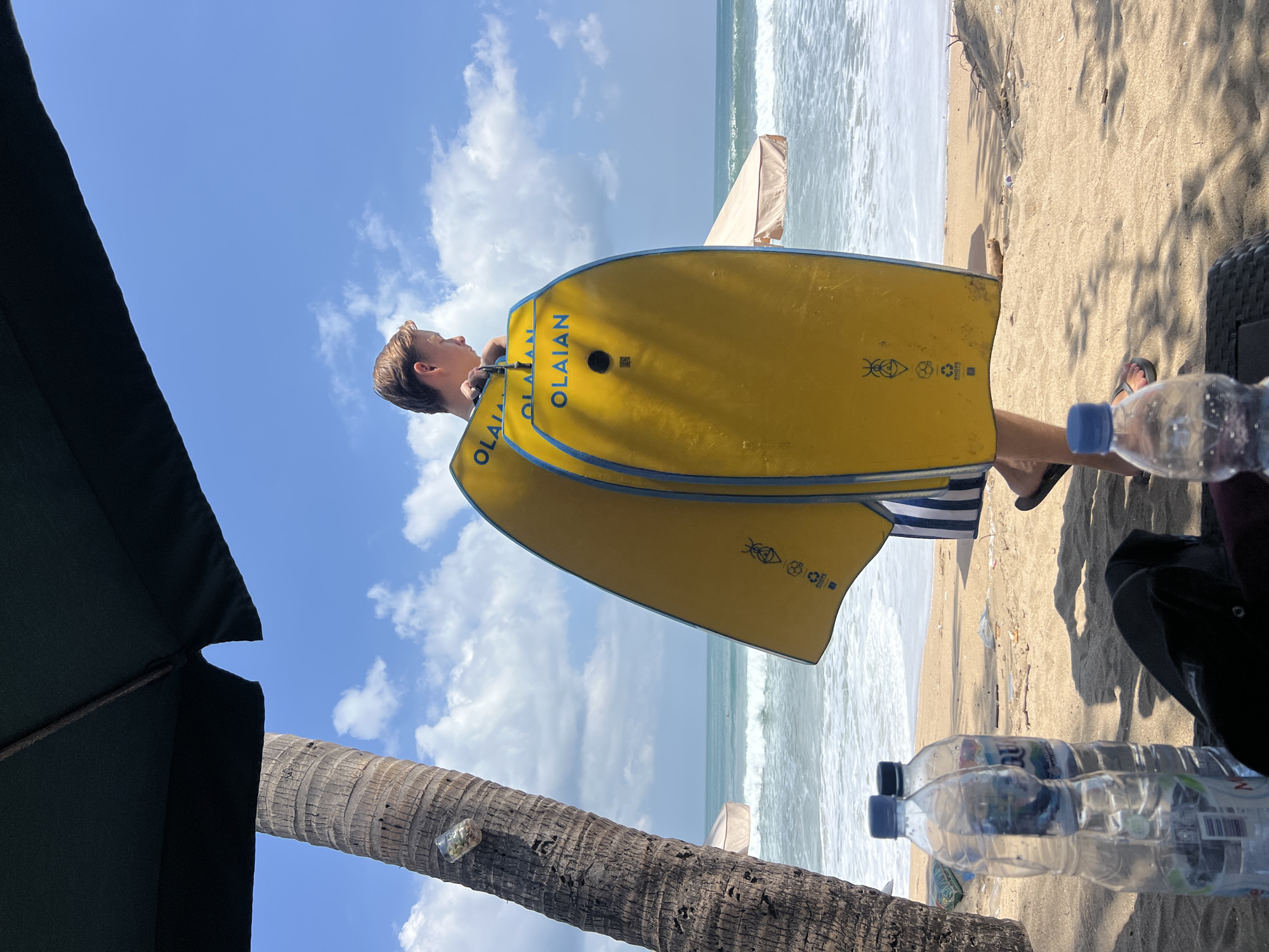 A young male stands on a sandy beach in Kuta, Indonesia, holding two yellow bodyboards with blue trim. He is partially obscured by an umbrella and a palm tree to his left. The ocean with gentle waves is visible behind him, under a bright blue sky with scattered clouds. In the foreground, two plastic water bottles sit on the sand, and a dark fabric object, possibly a bag or hat, is also present. Beach umbrellas and lounge chairs are visible further down the beach.
