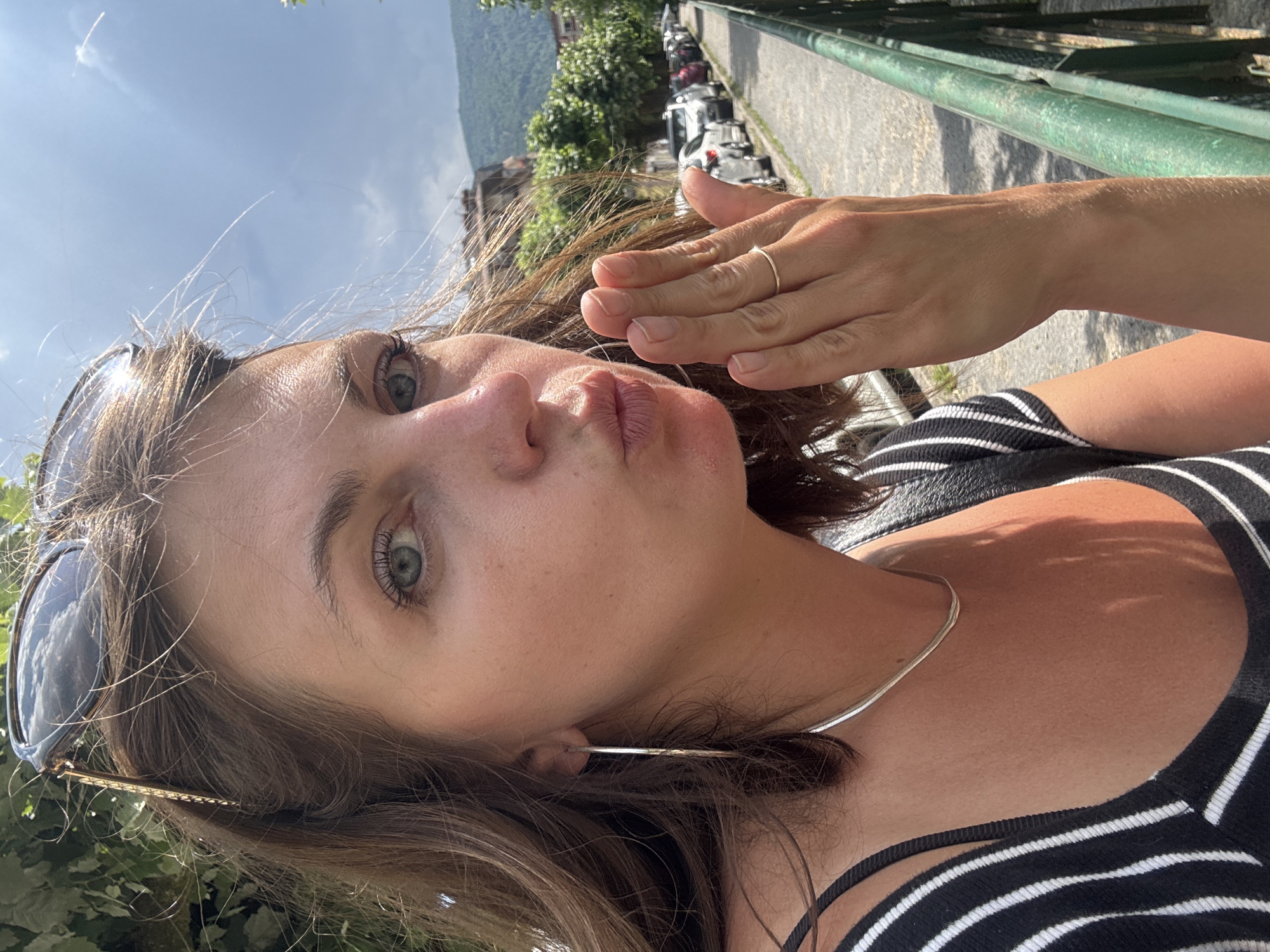 A woman with brown hair and blue-green eyes is taking a selfie outdoors, blowing a kiss. She is wearing a black and white striped top and a silver necklace. Her right hand is raised to her lips, and she is wearing a thin gold ring on her right ring finger. Sunglasses are perched on top of her head. The background shows a street lined with parked cars, trees, and buildings, suggesting an urban environment. The location is identified as Saint-Claude, France. The sky is partly cloudy.