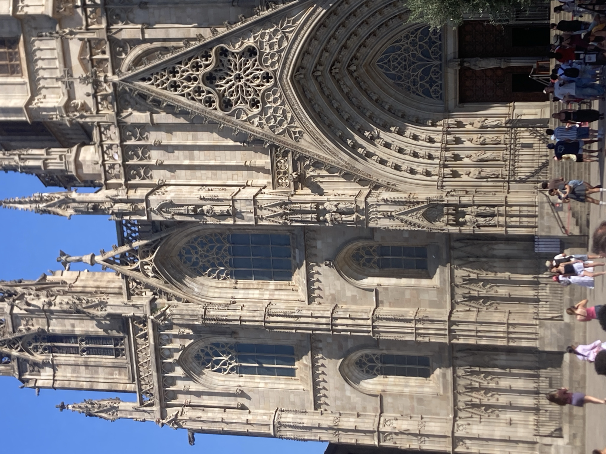 The image captures a vertical shot of the intricate facade of a large Gothic cathedral, likely in Barcelona, Spain, given the context. The architecture is elaborate, with detailed stonework, pointed arches, ornate tracery in windows, and numerous small statues and sculptural elements adorning the building. Several large arched windows with decorative patterns are visible, as are spires and smaller turrets projecting from the main structure. The building is made of light-colored stone, and the clear blue sky suggests it is daytime, likely sunny.
In the foreground and along the side of the cathedral, a number of people are visible, seemingly visiting or passing by. They are casually dressed, suggesting a tourist or public setting. Some are looking up at the building, while others are walking. The activity appears to be sightseeing or general pedestrian traffic in front of a significant landmark. The sunlight casts shadows on the facade, indicating the time of day. There is no visible text in the image.