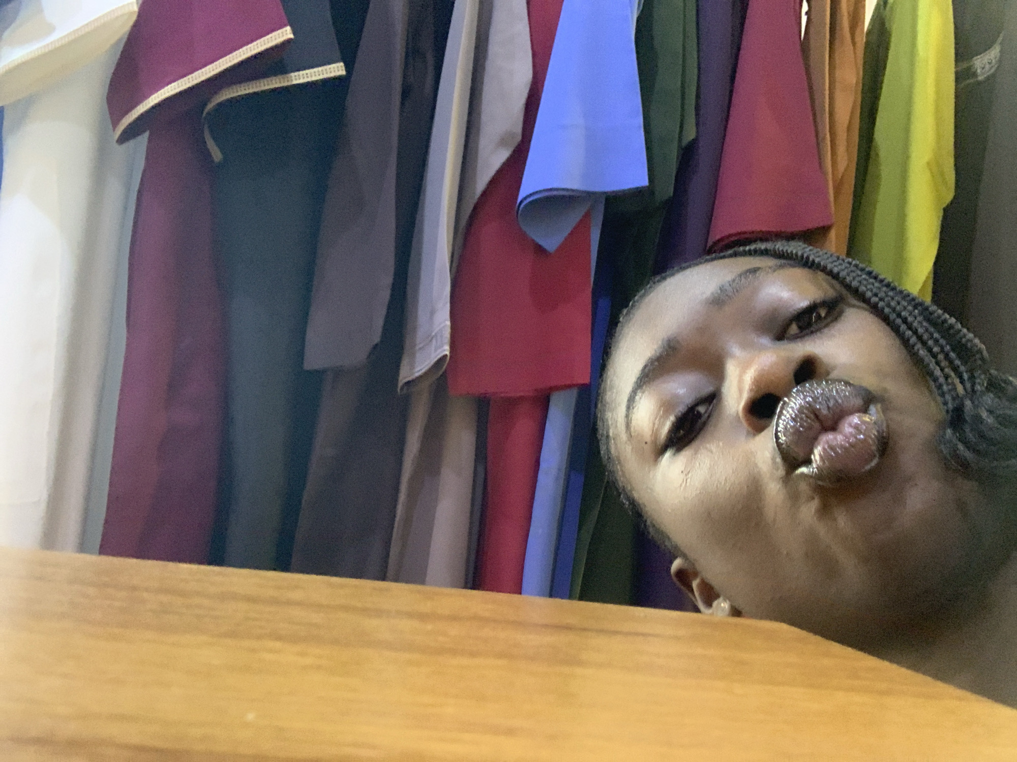 A close-up selfie shows a young Black person with braids puckering their lips, appearing to blow a kiss. The person is leaning over a wooden surface that occupies the foreground of the image. Behind them, a colorful array of fabrics hangs vertically, suggesting they may be in a tailor shop or a store selling textiles. The lighting is somewhat dim, with a slight glare on the person's lips. The background is a blur of clothing, in shades of white, maroon, dark gray, light gray, red, blue, green, purple, brown, and yellow. The image appears to have been taken indoors, likely in Gura Top, Nigeria, given the context.