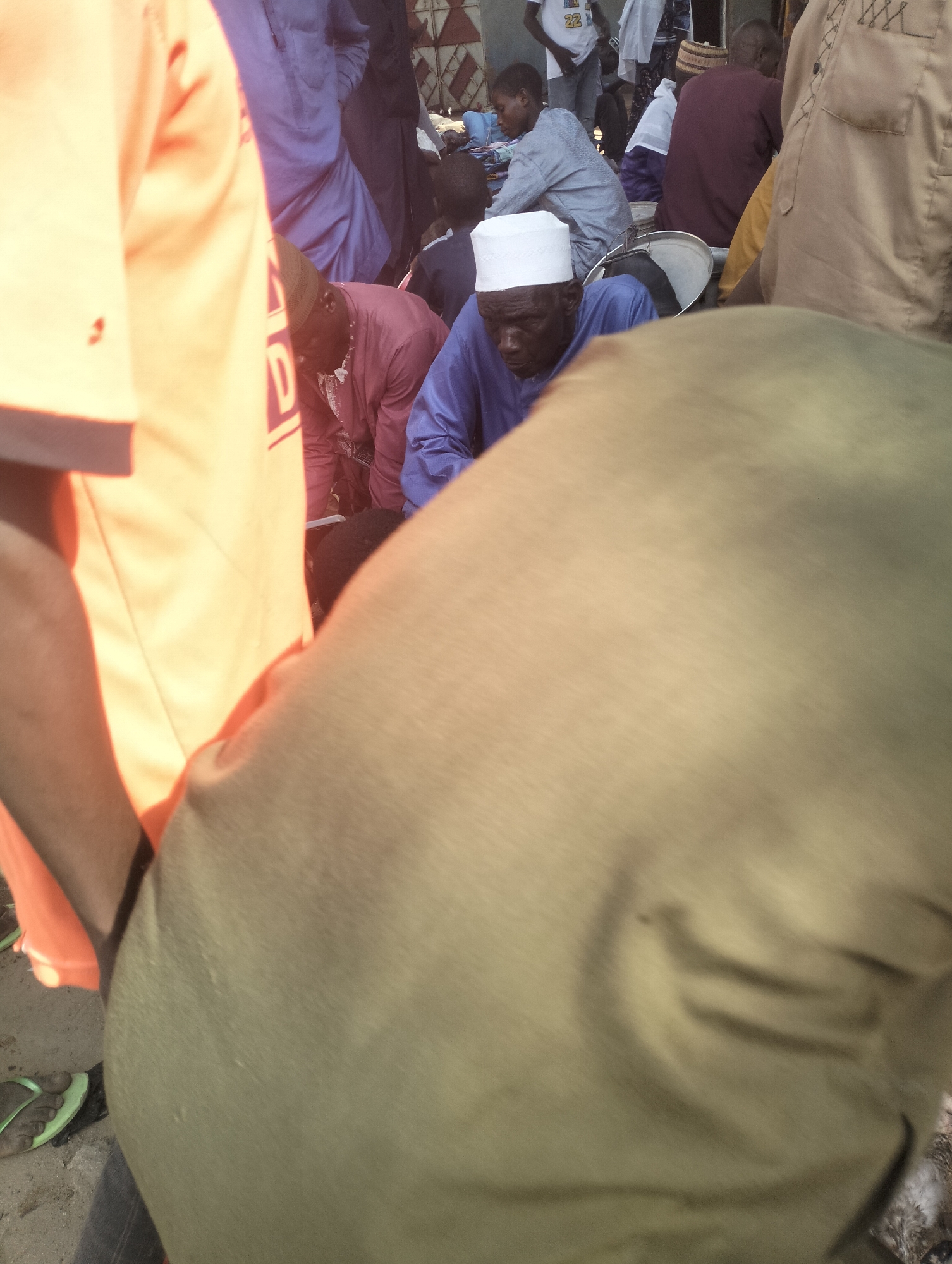 The image shows a crowded outdoor setting in the city of Unknown, Unknown. Several individuals are gathered, including a man in a blue robe and white cap. Other people are wearing various clothing, including orange shirts, purple robes, and casual wear. The scene appears to be a market or gathering of some kind, with metal trays and other objects visible amidst the crowd.