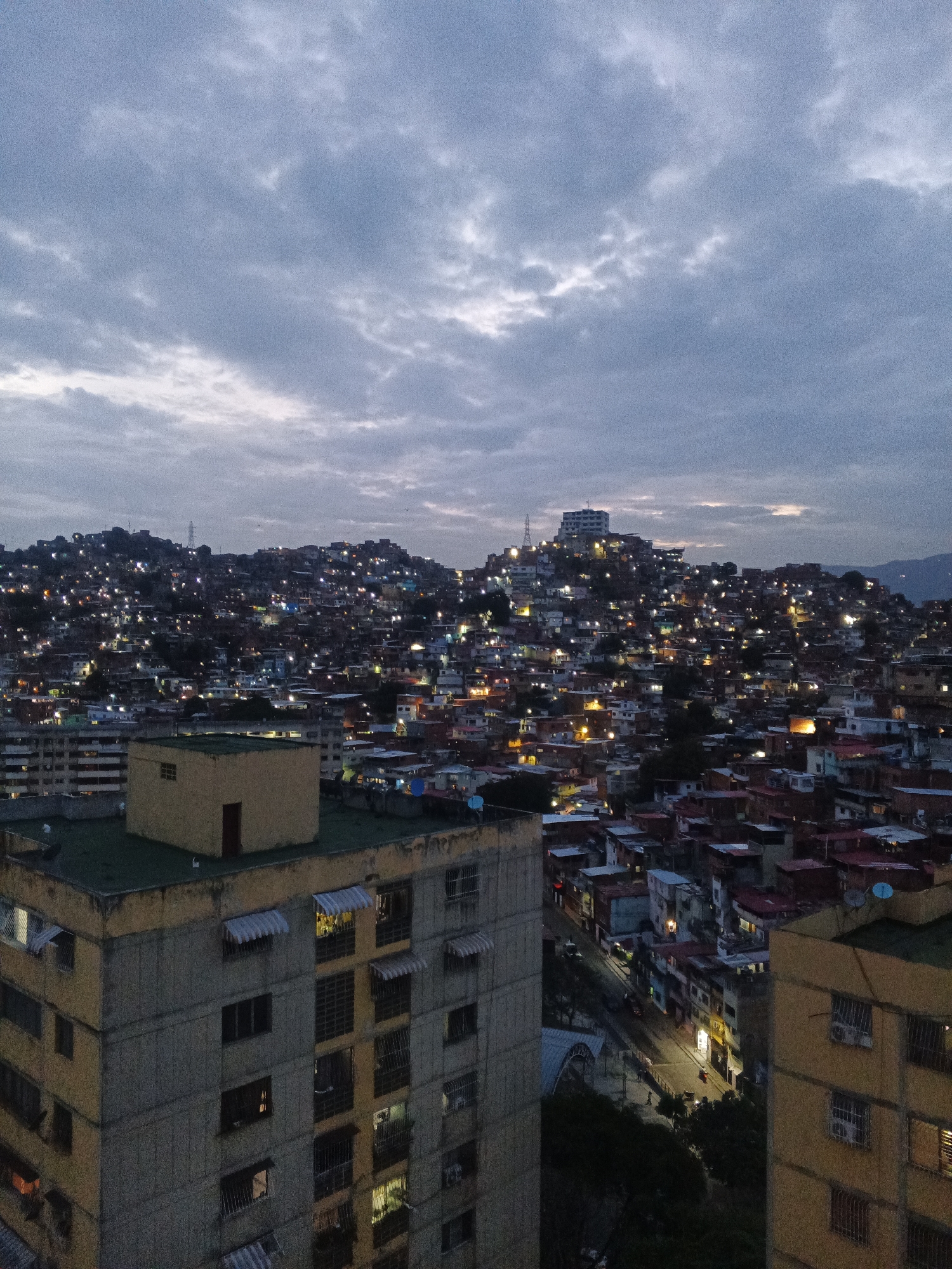 The image is an aerial, dusk shot of a densely populated hillside in Caracas, Venezuela, likely during the twilight hours. The foreground features the upper stories of several apartment buildings. The building on the left is light yellow with awnings over some windows. The building in the center is a smaller, tan structure with a dark door and a green roof. Another building, to the right, is yellow with visible air conditioning units. Beyond these structures, the hillside is covered with countless smaller buildings, many of which are illuminated by artificial lights, creating a speckled effect across the urban landscape. The sky above is overcast with grey clouds, tinged with the faint colors of sunset or sunrise. The overall impression is of a sprawling, vibrant, and illuminated city at nightfall.