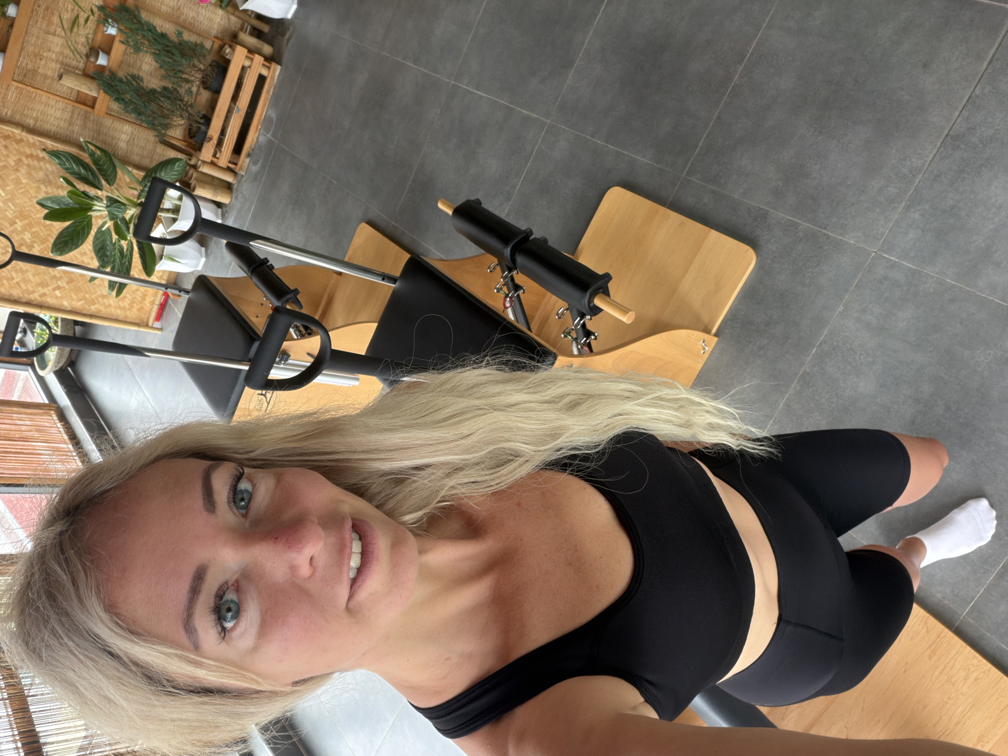 A woman with blonde hair and blue eyes is taking a selfie in a Pilates studio. She is wearing a black sports bra and black cycling shorts, with white socks on her feet. The studio has a wooden floor and grey tiled walls, with several Pilates reformers in the background. Plants are visible in the background, suggesting a bright and airy space. The overall impression is one of a fitness-focused environment. The location is not specified.