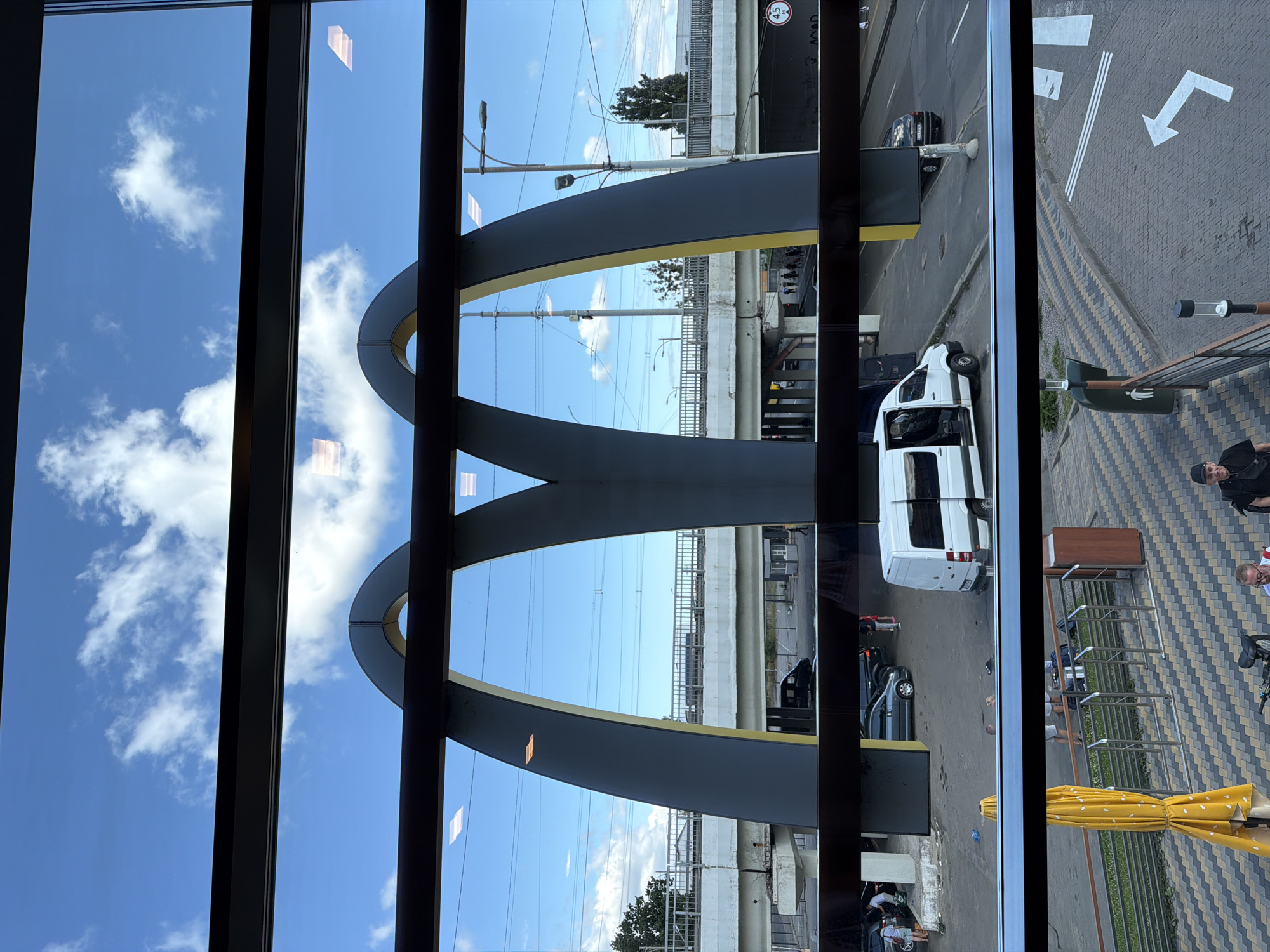 This image captures a view through a window with horizontal bars, revealing a McDonald's restaurant with its iconic golden arches. The scene appears to be in Kyiv, Ukraine, given the visible Cyrillic lettering on a traffic sign.
In the foreground, viewed from an elevated position, is a street with cars and pedestrians. A white van with its doors open is parked on the right side of the road. A black car is also visible. Further down, a person walks on a paved area, and a bicycle is leaning against a railing. Yellow and grey patterned paving stones are prominent.
Behind the McDonald's arches, a bridge spans across the road, with trains or trams visible on it. Power lines crisscross the sky. The sky is blue with scattered white clouds.
The overall impression is of a busy urban environment on a bright day. The McDonald's arches serve as a focal point, with the city infrastructure and daily life unfolding around it.