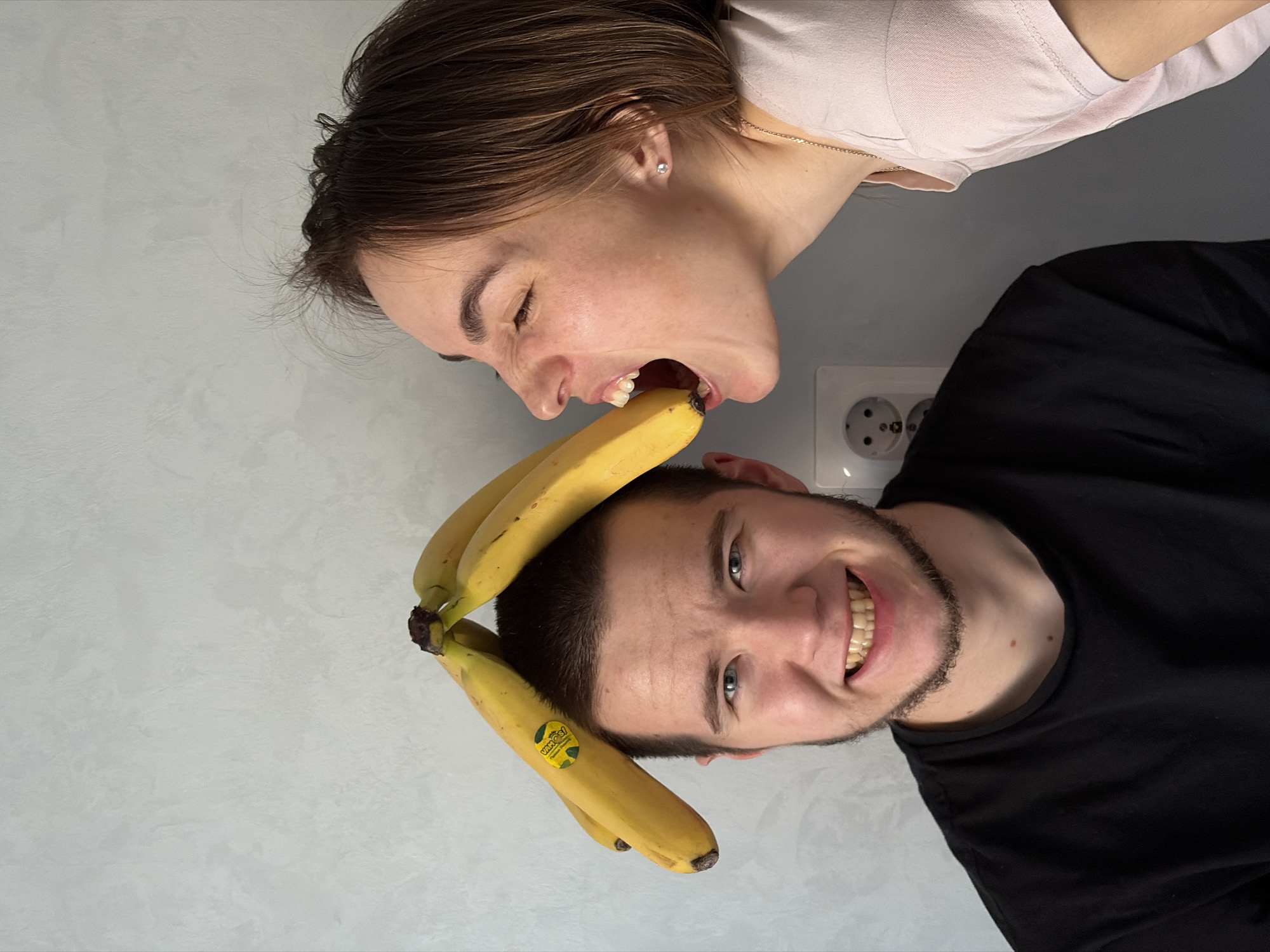 A young woman is attempting to bite a banana placed on top of a young man's head. The woman is wearing a light pink shirt and the man is wearing a black shirt. The background is a gray wall with a white electrical outlet. The image appears to be rotated approximately 90 degrees counterclockwise. It is not possible to verify the location in Sarapul, Russia from this image.