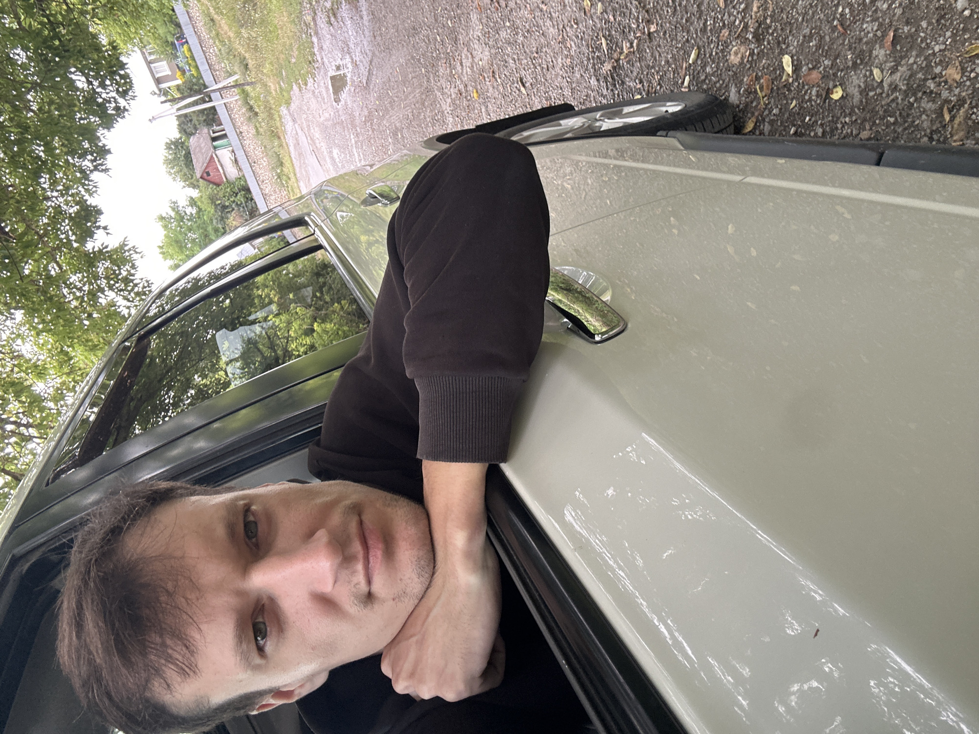 A man is leaning his arms on the open window of a light-colored SUV, looking out at the viewer. He has brown hair and a light complexion, with a slight mustache and stubble. He is wearing a dark hoodie. The car is parked on a gravel road, with a small puddle of water visible. In the background, there are trees, a stone wall, and a few small houses with red roofs. The overall atmosphere is overcast and slightly damp. The location appears to be a rural area, likely a village or outskirts of a town.