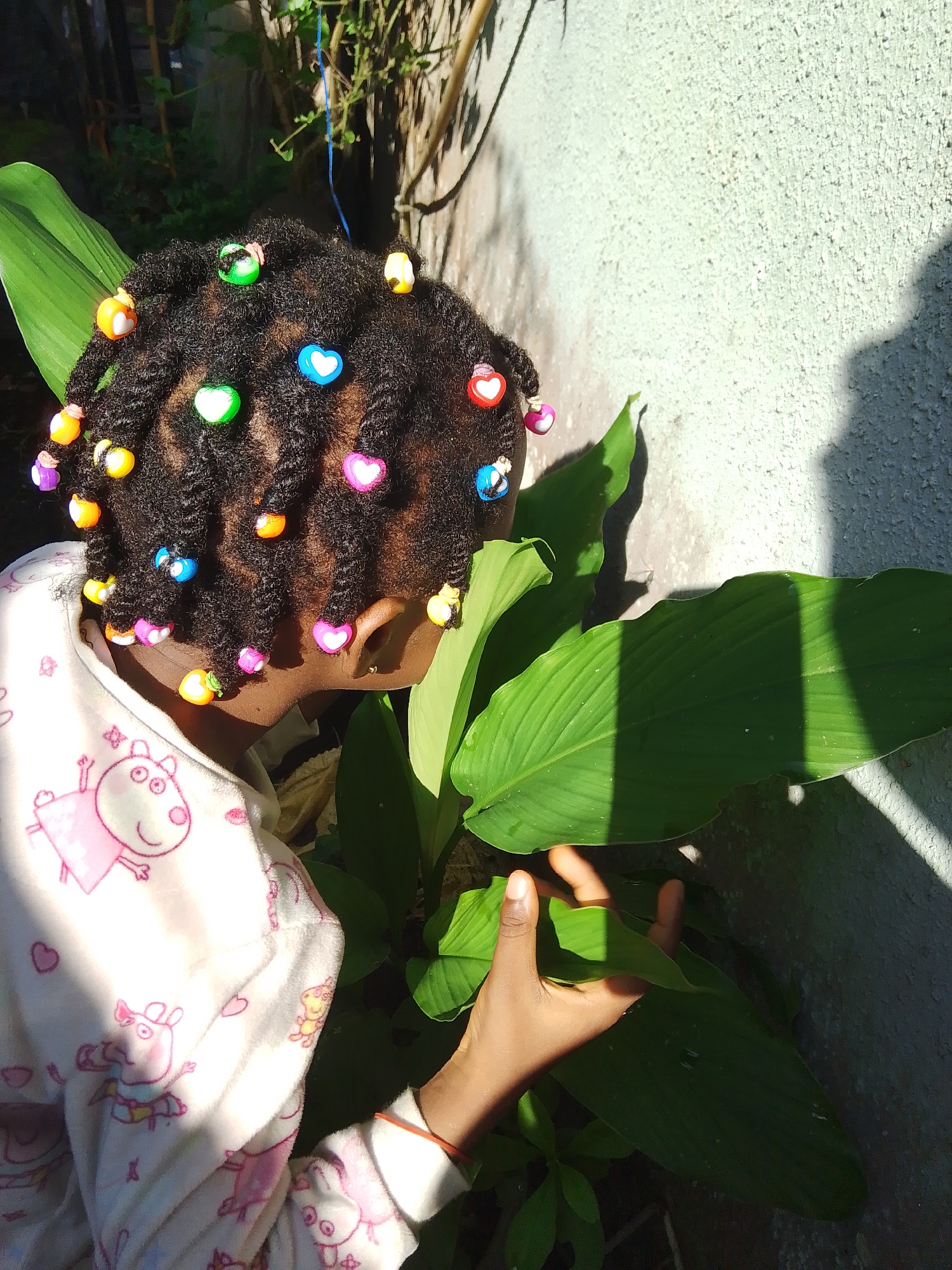 In an outdoor urban environment, a young Black child with braided hair adorned with colorful beads, including heart shapes and other charms, is partially visible from the side. The child is wearing a white fleece robe with a pattern of pink cartoon characters, likely from the show Peppa Pig. The child's hand is reaching towards large green leaves of a plant. The background consists of a textured, light-colored wall and some shadowy foliage, suggesting it is daytime. The lighting indicates direct sunlight, casting shadows on the wall and leaves. The specific location is Unknown, Unknown.