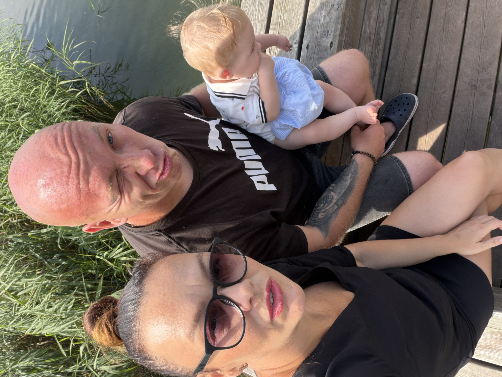 A selfie shows a family of three on a wooden pier by a body of water surrounded by tall green reeds. A woman with her hair in a bun and wearing sunglasses is in the foreground, on the left, looking at the camera. Next to her is a bald man winking at the camera. Between the man and the woman, a baby with blonde hair is being held. The baby is wearing a white and blue shirt and light blue shorts. The man is wearing a black Puma t-shirt and dark shorts. The woman is wearing a black v-neck dress. The scene appears to be outdoors on a sunny day.