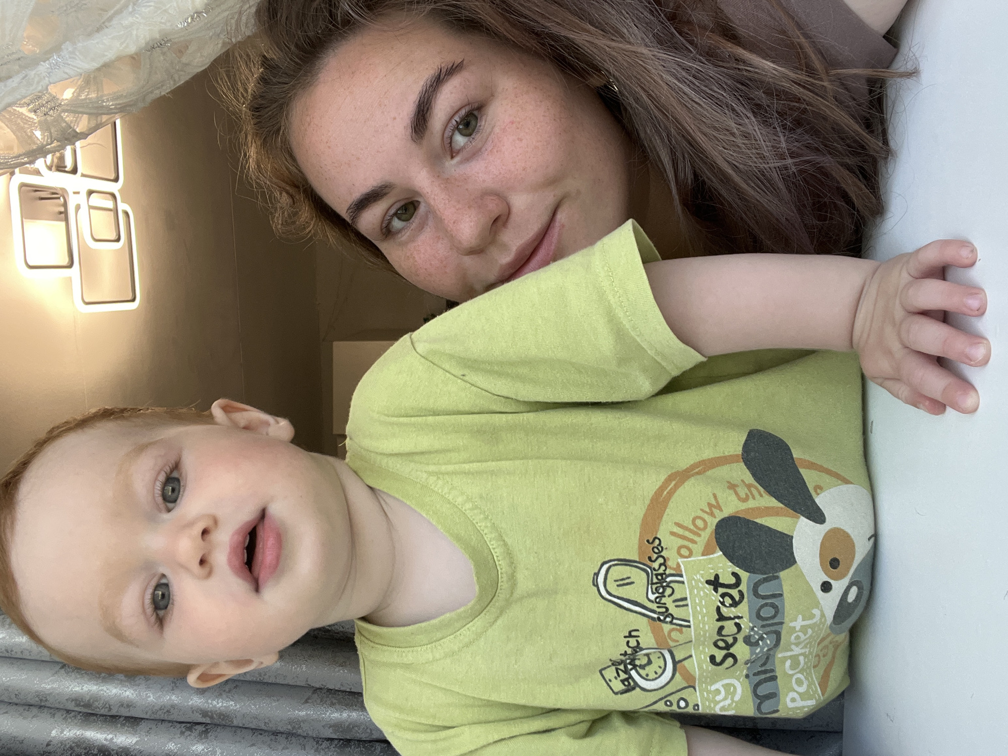 This is a medium shot, eye-level, indoor photograph featuring a baby and a woman. The baby, positioned on the left, has fair skin and reddish hair. The woman is on the right, with her face turned towards the camera and a visible smile. She has light brown hair and a smattering of freckles across her face. The baby is wearing a light green t-shirt with a cartoon print. The background shows a ceiling light fixture and curtains. The overall impression is of a candid, domestic moment. The image does not provide any specific information that would allow for the identification of the location within the city of Chaikovsky, Russia.