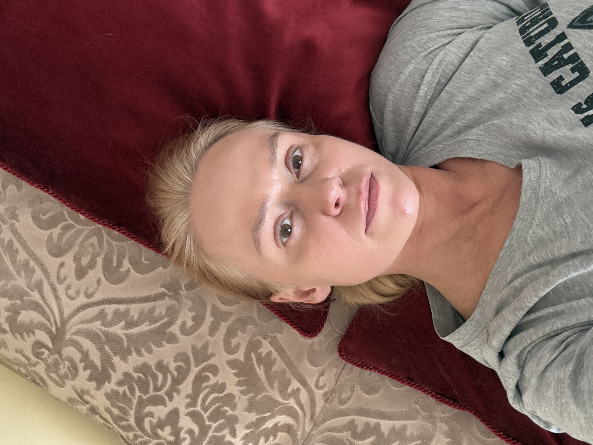 A fair-skinned woman with blonde hair is lying on a patterned pillow next to a red pillow. She is wearing a gray t-shirt with black letters. The location in the city of Chaikovsky, Russia cannot be determined from this image.