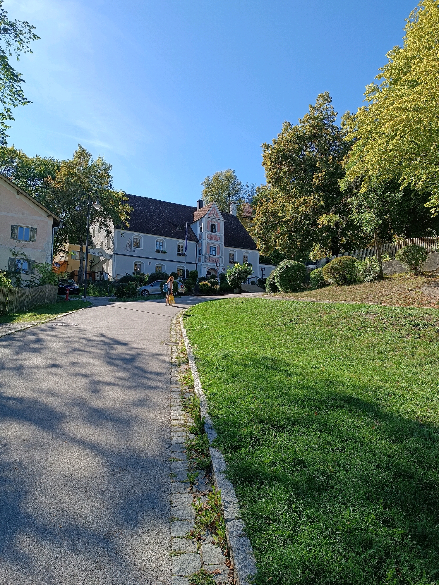 This image depicts a sunny day scene in the town of Andechs, Germany. In the foreground, an asphalt road curves to the right, bordered by a stone curb and a vibrant green lawn. Shadows of trees are cast across the road. A woman with a yellow bag and backpack is walking on the side of the road. Further back, several buildings are visible, including a large, light-blue building with a dark roof, adorned with architectural details and signage. Adjacent to it is a lighter-colored building. Mature trees with green and yellow foliage line the background, suggesting an autumn or late summer setting. The sky is clear and bright blue. The overall impression is one of a serene, well-maintained outdoor environment.