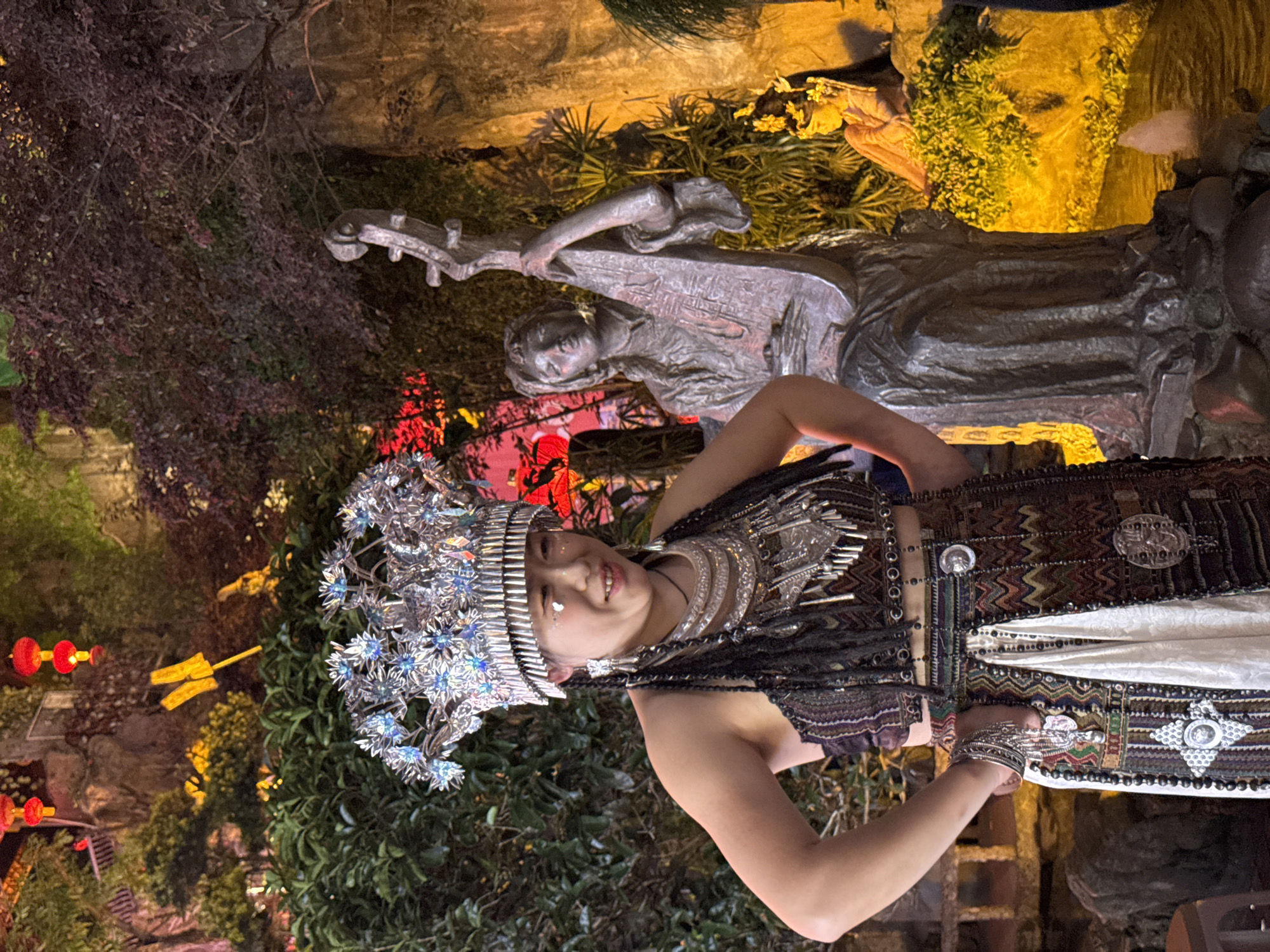 A woman in traditional clothing and an elaborate headdress poses in front of a statue of a musician in the Yuzhong District of China. The headdress is silver with blue accents. Her outfit is colorful and features geometric patterns. A statue of a woman reclining and holding a stringed instrument is behind her. Foliage, lighting, and stone structures surround the subject and statue. Red lanterns hang in the background.