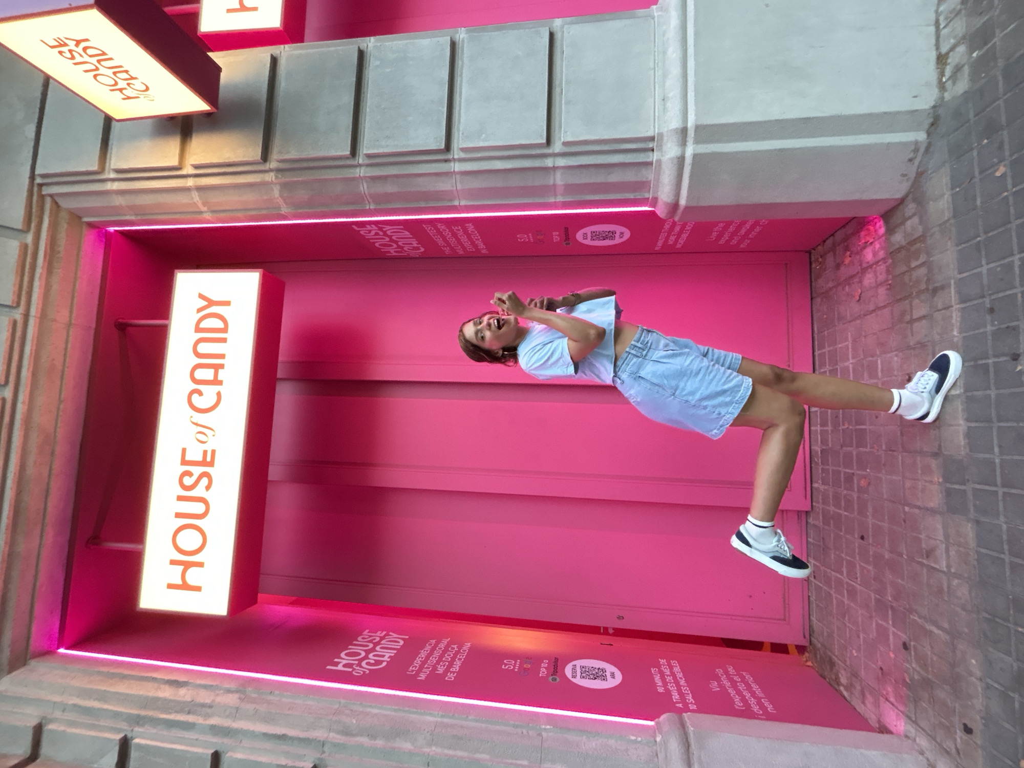 A young woman with dark hair, wearing a light blue crop top and denim shorts, is captured mid-action outside a vibrant pink establishment. She is playfully leaning back, with one leg extended, and her mouth is open as she holds a pink lollipop to her lips, seemingly about to take a bite. The setting is clearly urban, indicated by the tiled pavement and the architectural elements of the building. A bright, illuminated sign above the entrance reads "HOUSE of CANDY" in orange lettering, with a smaller sign below also bearing the same name. The pink facade of the building is accentuated by neon pink lighting. Text on the wall and floor further identifies the location as "HOUSE of CANDY" and describes it as a "SWEETEST MULTISENSORY EXPERIENCE IN BARCELONA". Additional text in Catalan mentions "L'EXPERIÈNCIA MULTISENSORIAL MÉS DOLÇA DE BARCELONA" and "Viu l'experiència i desperta el teu nen interior!". Other visible text indicates a "5.0" rating and mentions "Tripadvisor" and the duration of the experience as "90 MINUTS" through "10 SALES INCREÍBLES". The overall impression is one of fun and excitement, likely during daytime given the bright lighting.