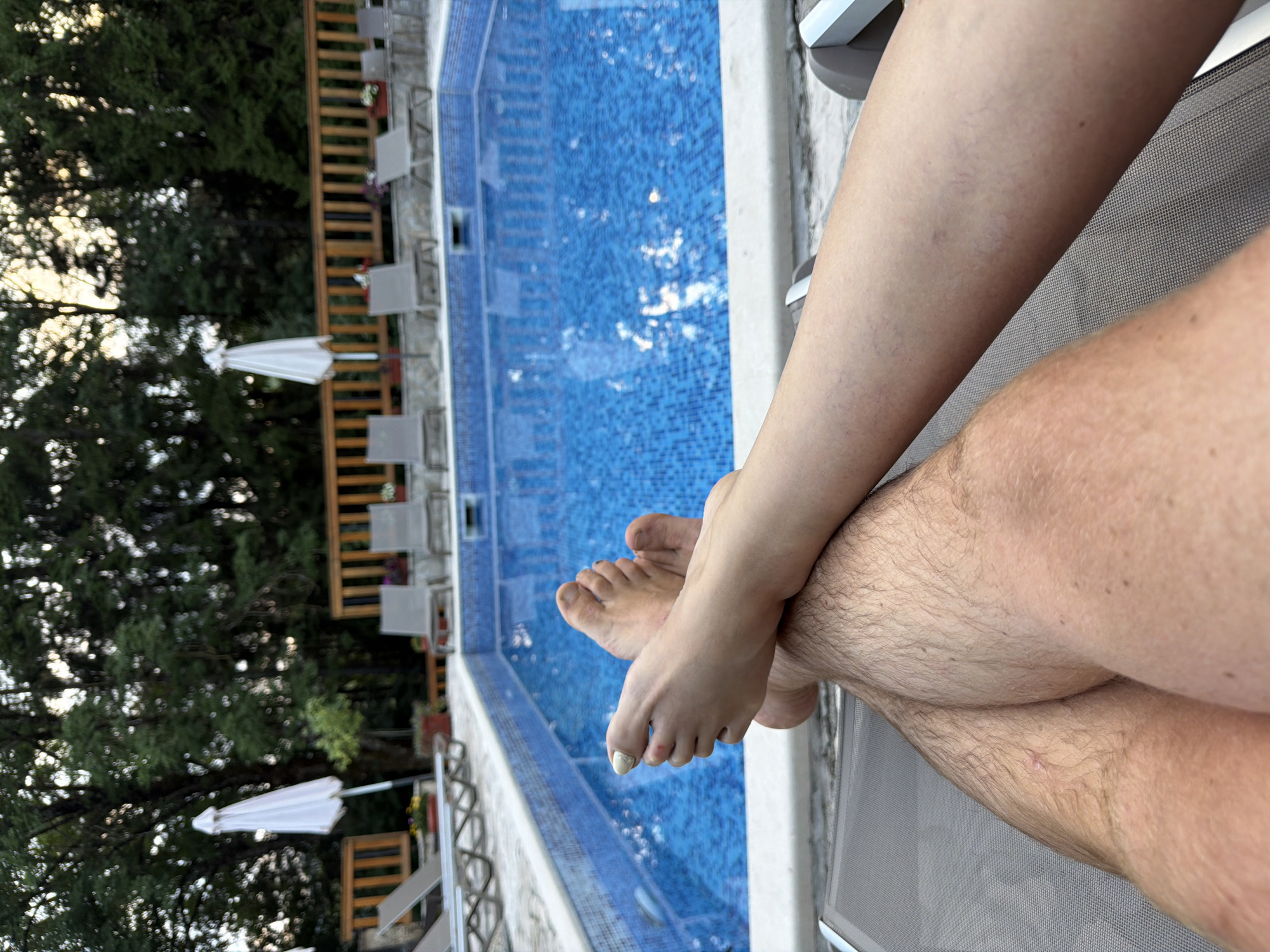 The image shows a close-up view of two pairs of legs and feet resting on lounge chairs by a swimming pool. One pair of legs belongs to a person with hairy legs, and the other to a person with smooth, lightly tanned skin. The feet are crossed, with one foot adorned with light green nail polish. The swimming pool is filled with blue water and is surrounded by a white tiled edge. In the background, there are several empty lounge chairs and umbrellas, as well as a wooden fence and lush green trees. The scene appears to be an outdoor relaxation area, likely a resort or hotel, given the organized setup of the pool and loungers. The lighting suggests it is either early morning or late afternoon. The location is stated as Kolašin, Montenegro, suggesting this is a scene from a resort or private property within that city, known for its natural beauty and outdoor activities.
